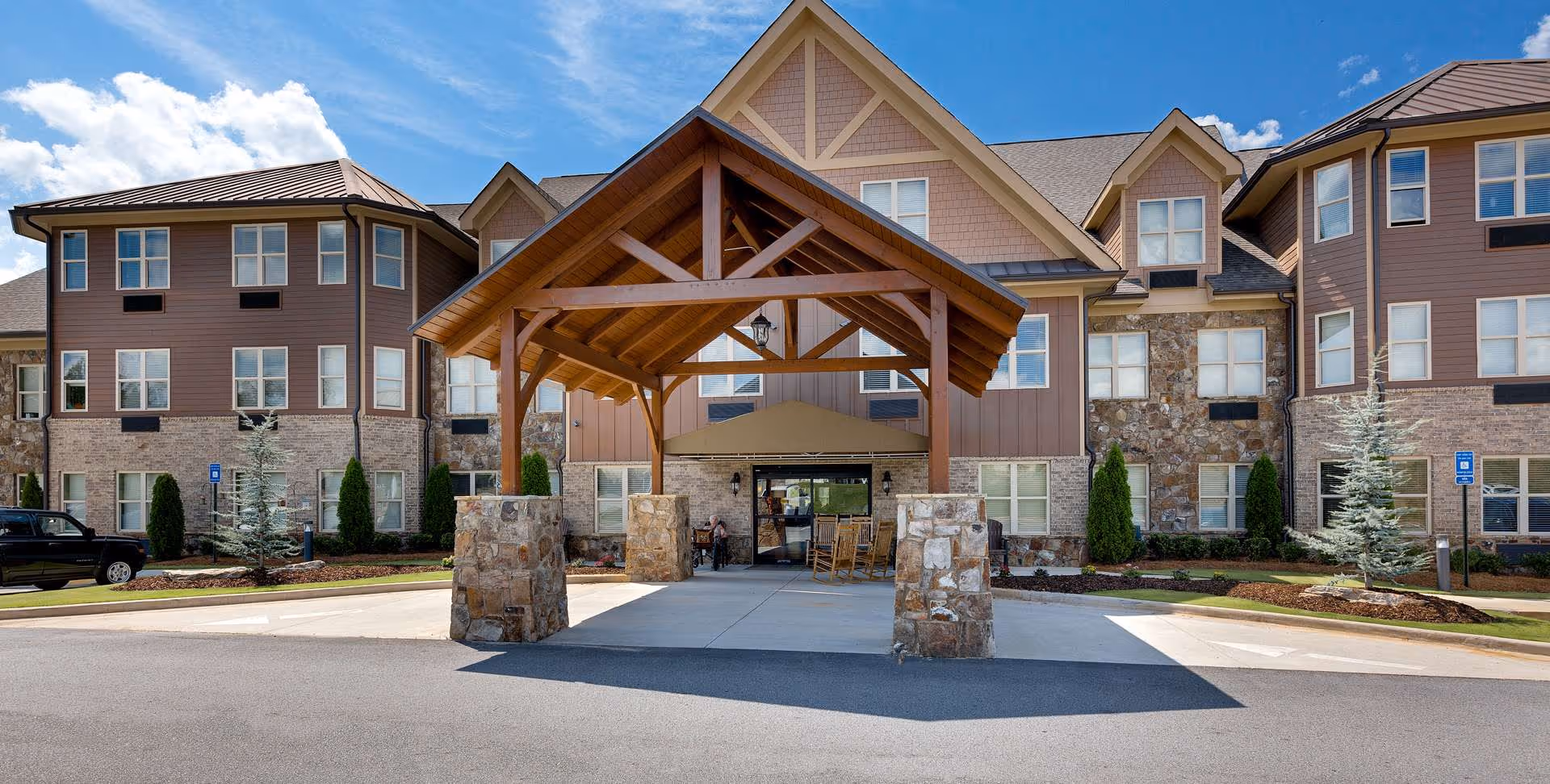 Front exterior view of Oaks at Acworth senior living facility showing a large covered entrance with wooden beams and stone pillars, a paved driveway, landscaped greenery, and a multi-story building with multiple windows under a blue sky with some clouds.