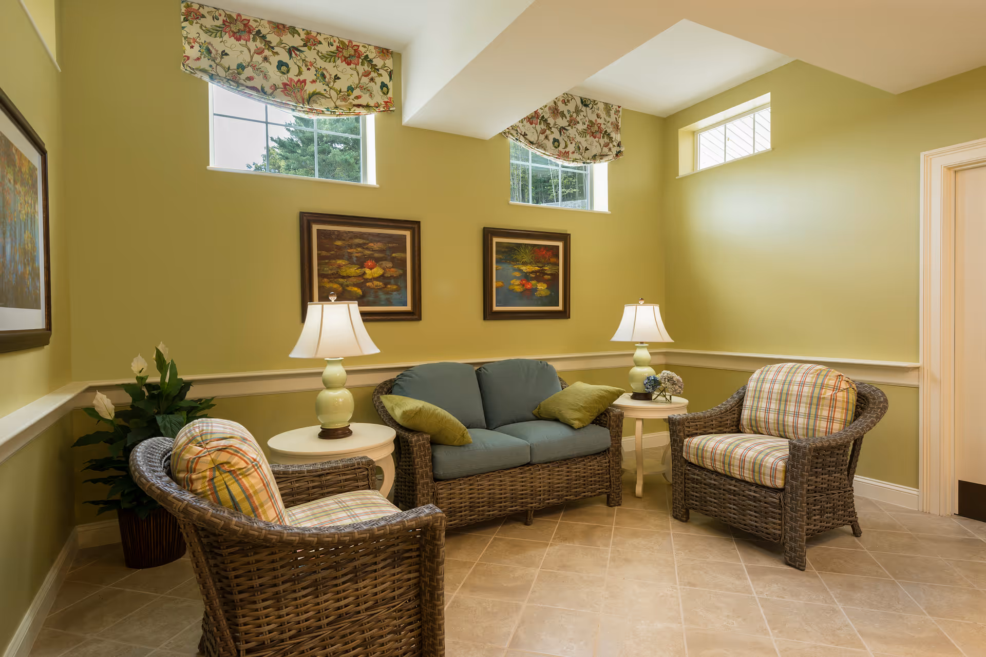A cozy sitting area with green walls featuring two small windows with floral valances. The room contains a wicker loveseat with blue cushions and two matching wicker armchairs with plaid cushions. There are two white side tables each holding a lamp with a white shade. Two framed paintings of water lilies hang on the wall above the loveseat. A potted plant is placed in the corner.