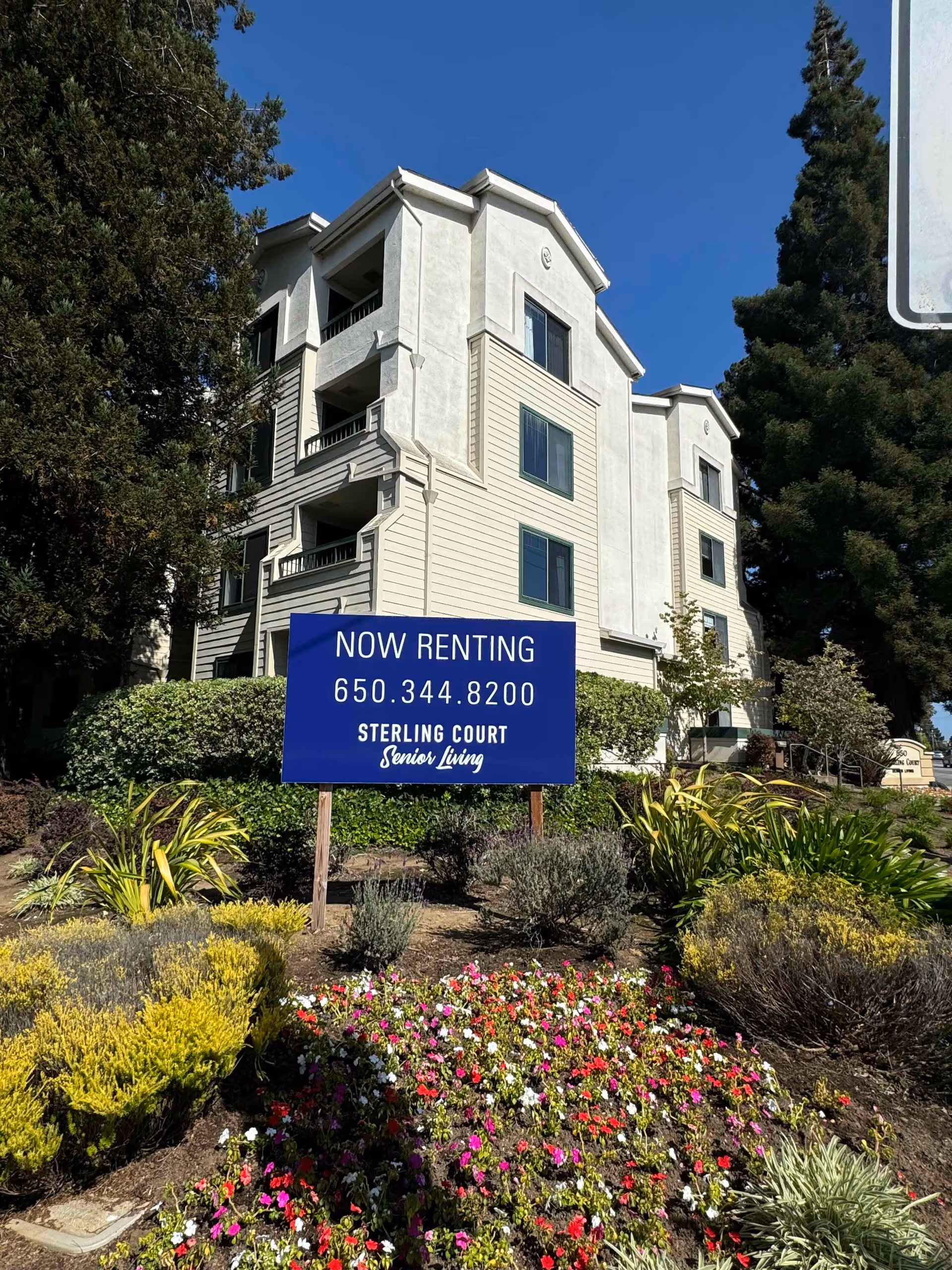 Exterior front view of a multi-story senior living building with landscaping and a blue 'Now Renting - Sterling Court Senior Living' sign showing a phone number.