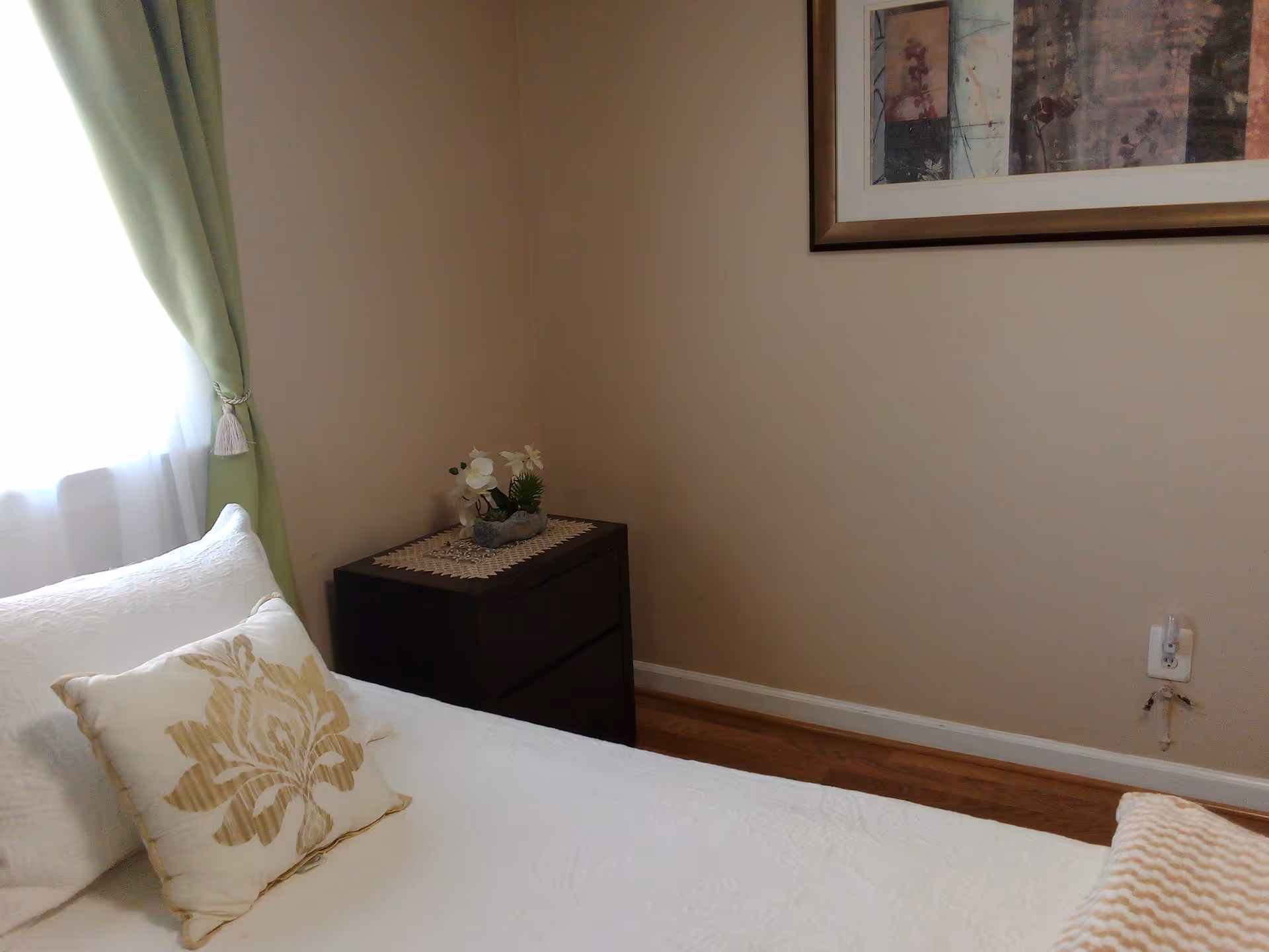 A tidy bedroom with a bed, nightstand, framed art, and green-curtained window.