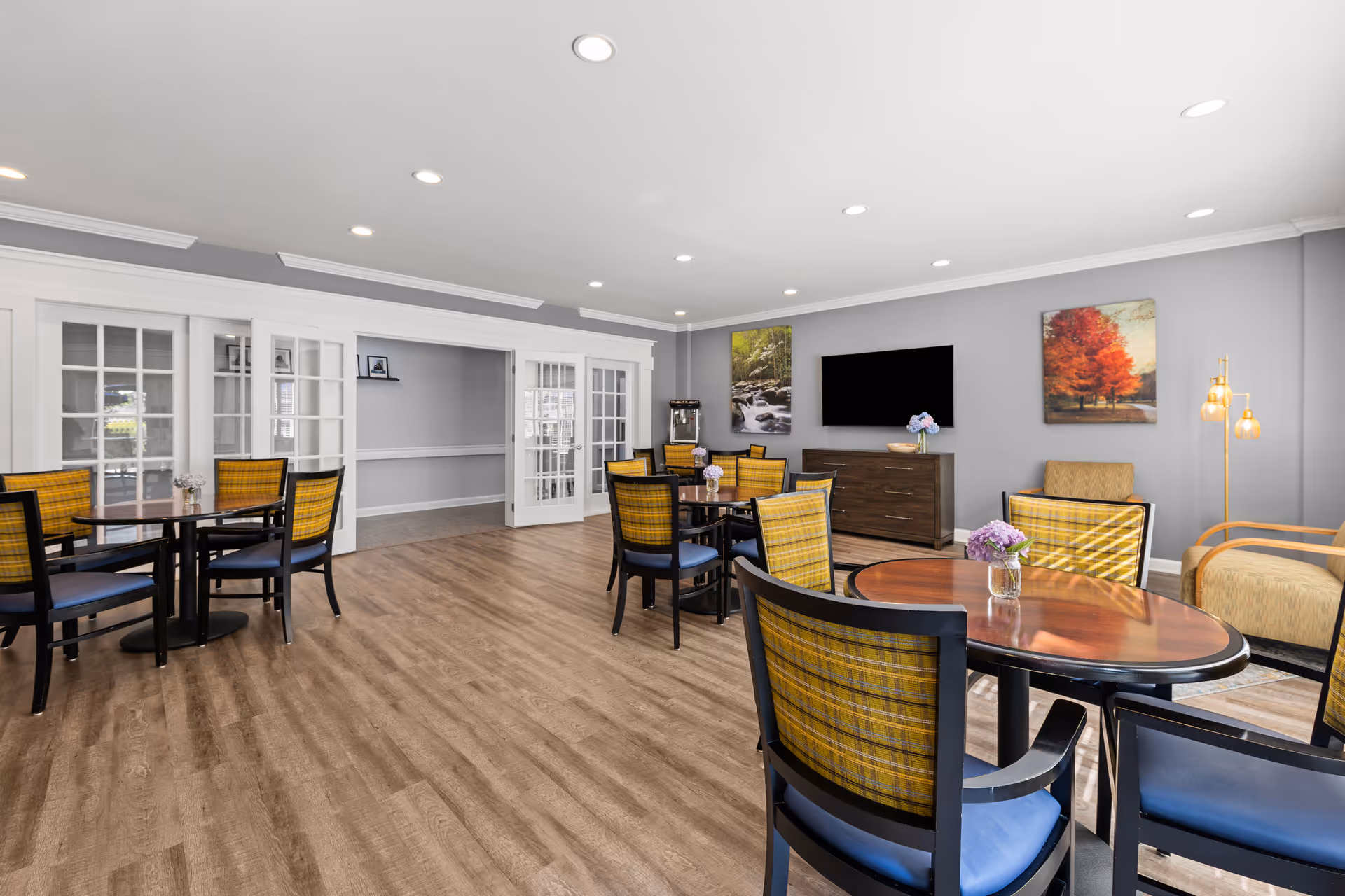 A spacious, well-lit common area with multiple round tables and chairs featuring yellow plaid backs and blue seats. The room has wood flooring, gray walls, and recessed ceiling lights. There is a flat-screen TV mounted on the wall above a wooden cabinet, two landscape paintings, a floor lamp with three bulbs, and a water dispenser in the corner. French doors lead to an adjacent room.