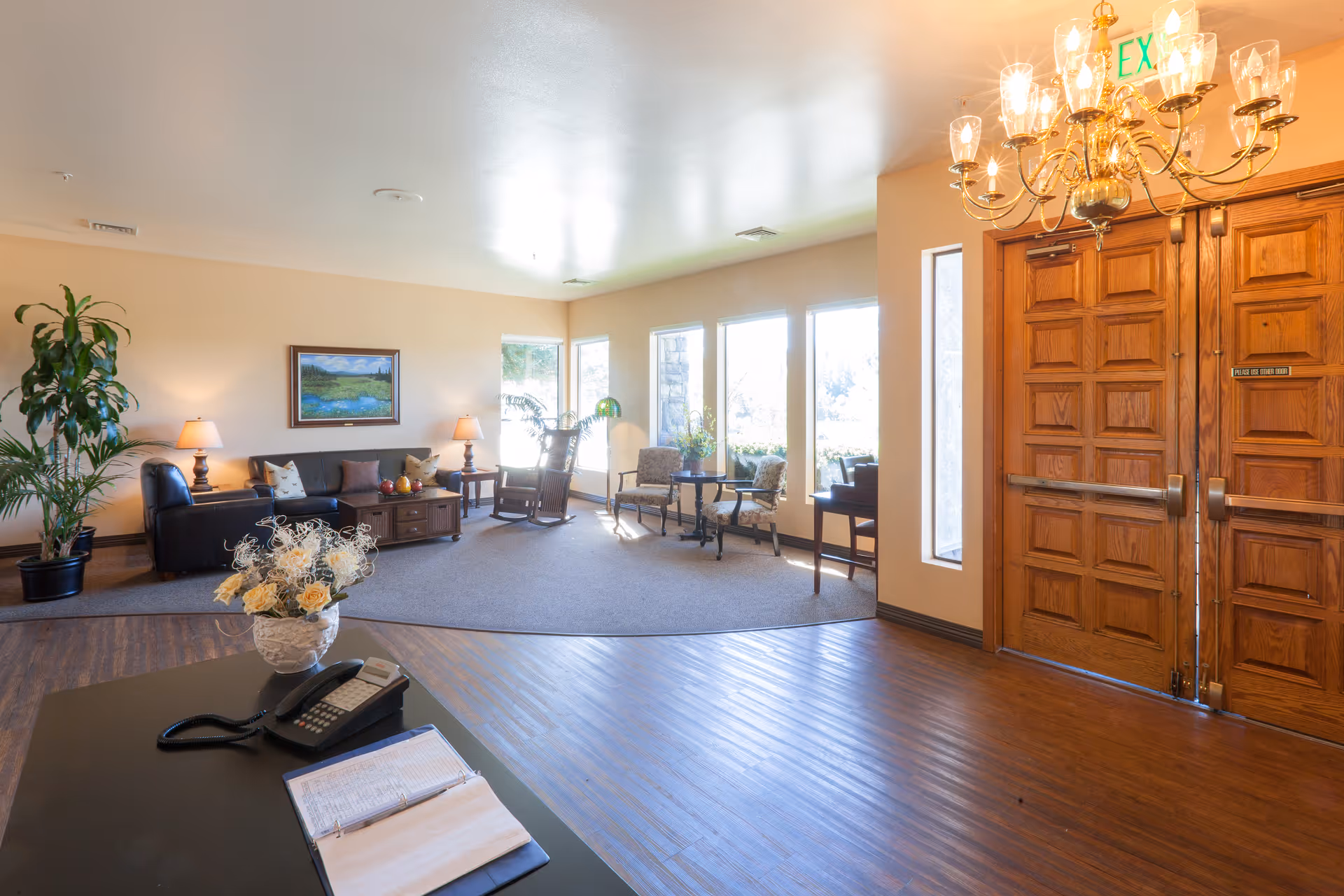 A bright and welcoming common area with wooden double doors, a chandelier, a seating area with a black leather couch, chairs, a coffee table, and large windows letting in natural light. There is a desk with a phone and an open binder in the foreground, and a potted plant and wall art in the background.