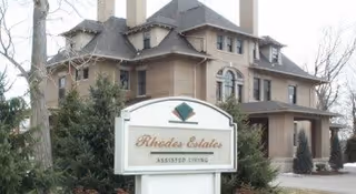 Brick multi-story assisted living building with a front sign that reads "Rhodes Estates Assisted Living" and trees in the foreground.