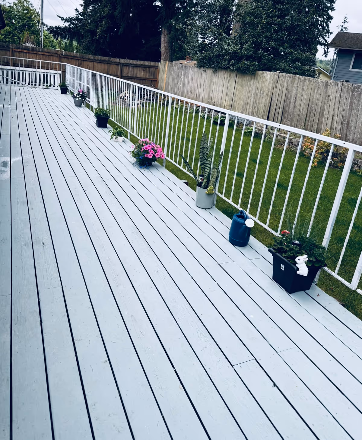 Light gray wooden outdoor deck with potted plants along a white railing and green lawn.