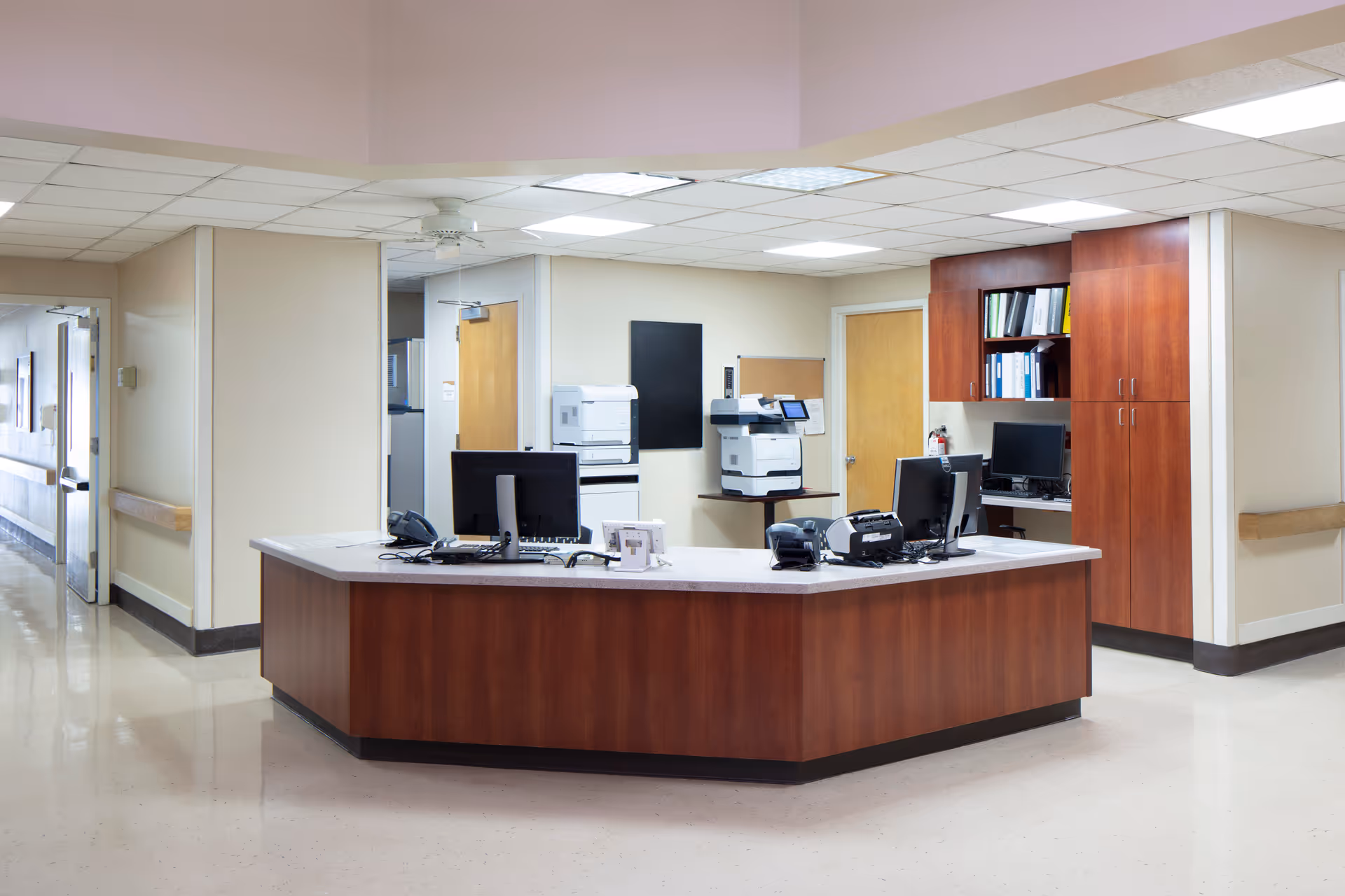 Interior reception area featuring a curved wooden desk, computers, printers, and storage cabinets.