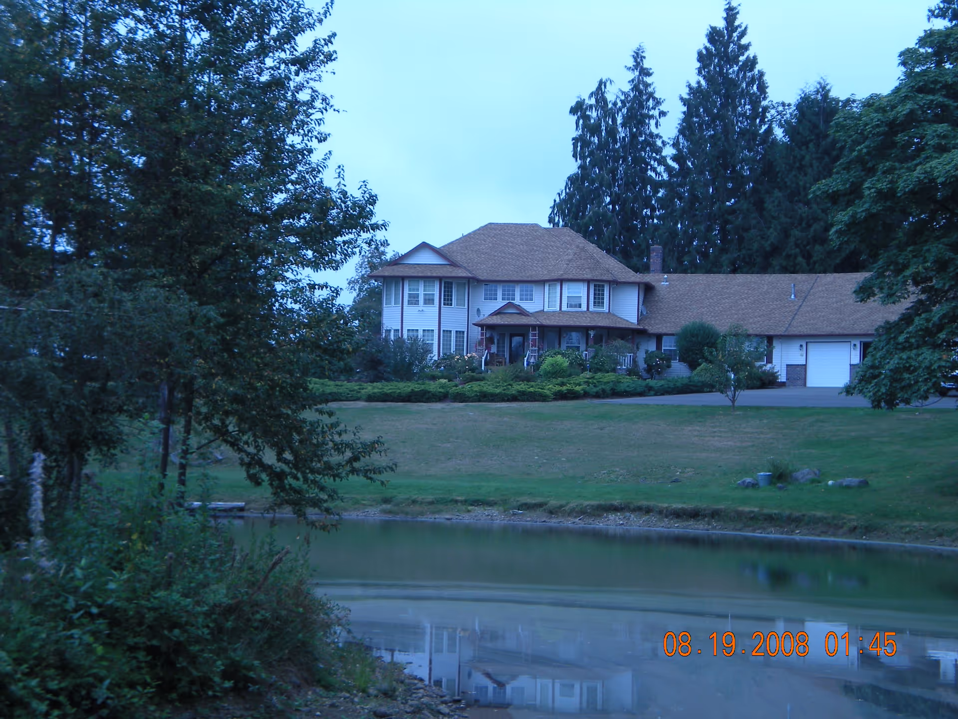 Two-story house with a large lawn and a pond in the foreground, surrounded by trees.