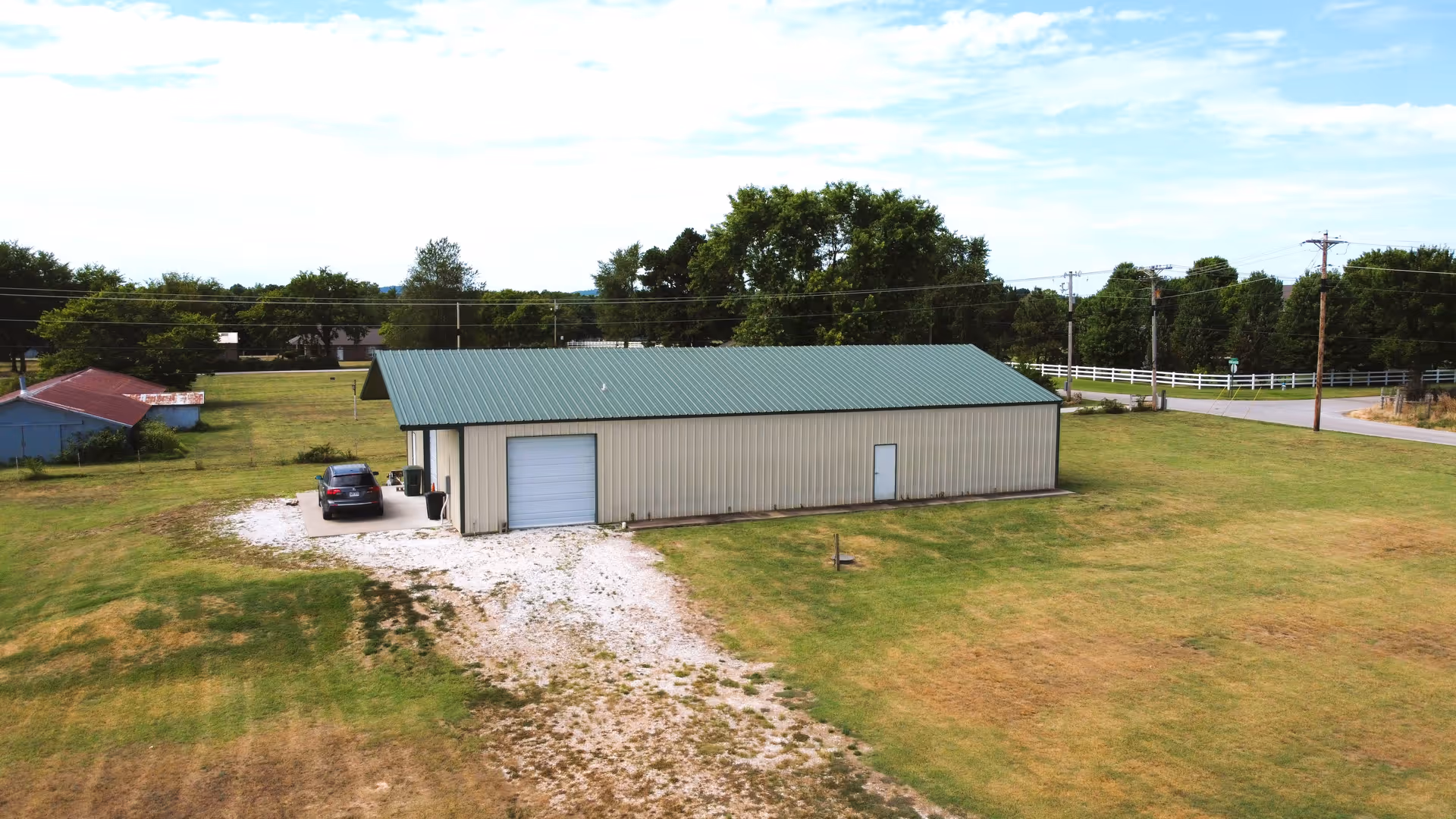 Metal-roofed rectangular building sits on grassy rural property with a gravel driveway and a car.