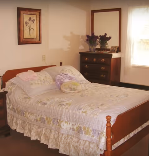 A cozy bedroom with a wooden bed frame and a floral quilted bedspread. The bed has several pillows and a small decorative cushion. To the right of the bed is a wooden dresser with a large mirror, a vase of purple flowers, and a framed photo. A window with sheer white curtains allows natural light into the room. A framed floral artwork hangs on the wall above the bed.