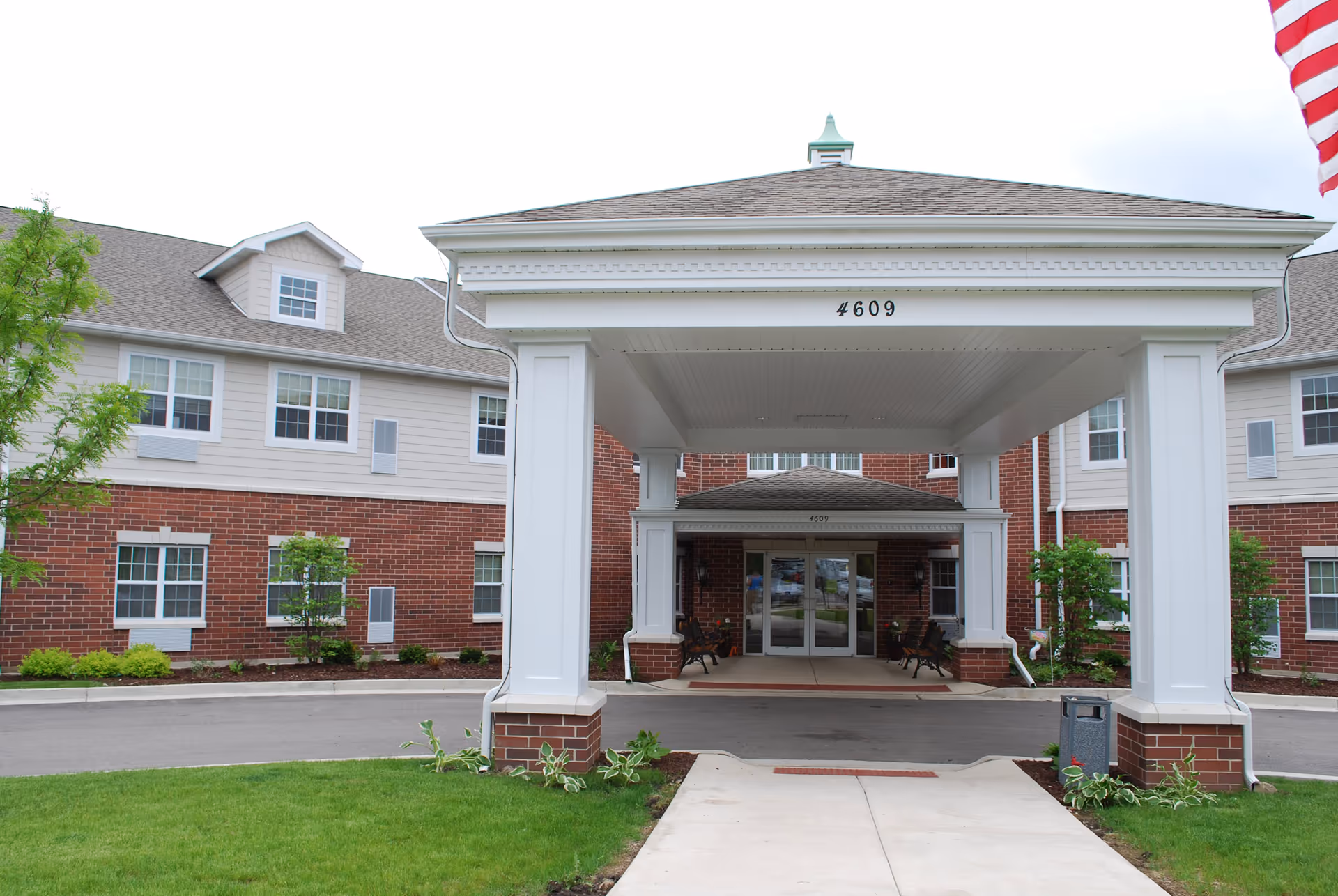 Entrance of a senior living facility with a covered drop-off area supported by white columns. The building has a combination of red brick and light-colored siding with multiple windows. The address number 4609 is displayed on the front of the covered entrance. There is a small landscaped area with green grass and plants, and an American flag is partially visible on the right side.