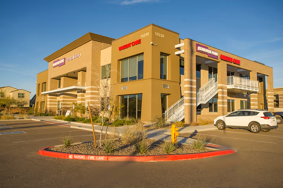Two-story tan medical building with urgent care signs and a parking lot in front.