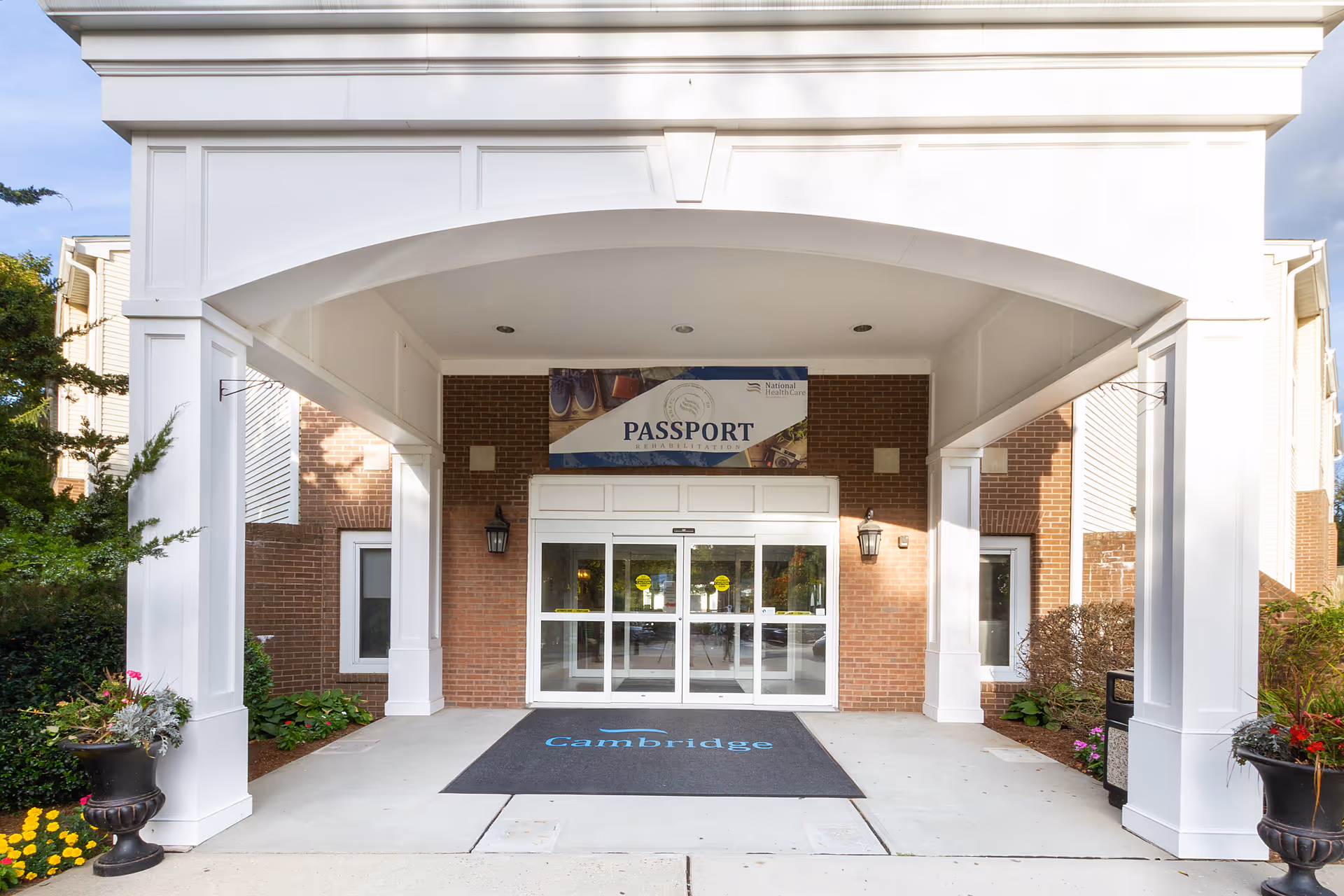 Entrance to Cambridge Health and Rehabilitation Center featuring a covered walkway with white pillars, a black mat with the word 'Cambridge' in blue, glass double doors, brick walls, and a sign above the doors that reads 'PASSPORT Rehabilitation'.