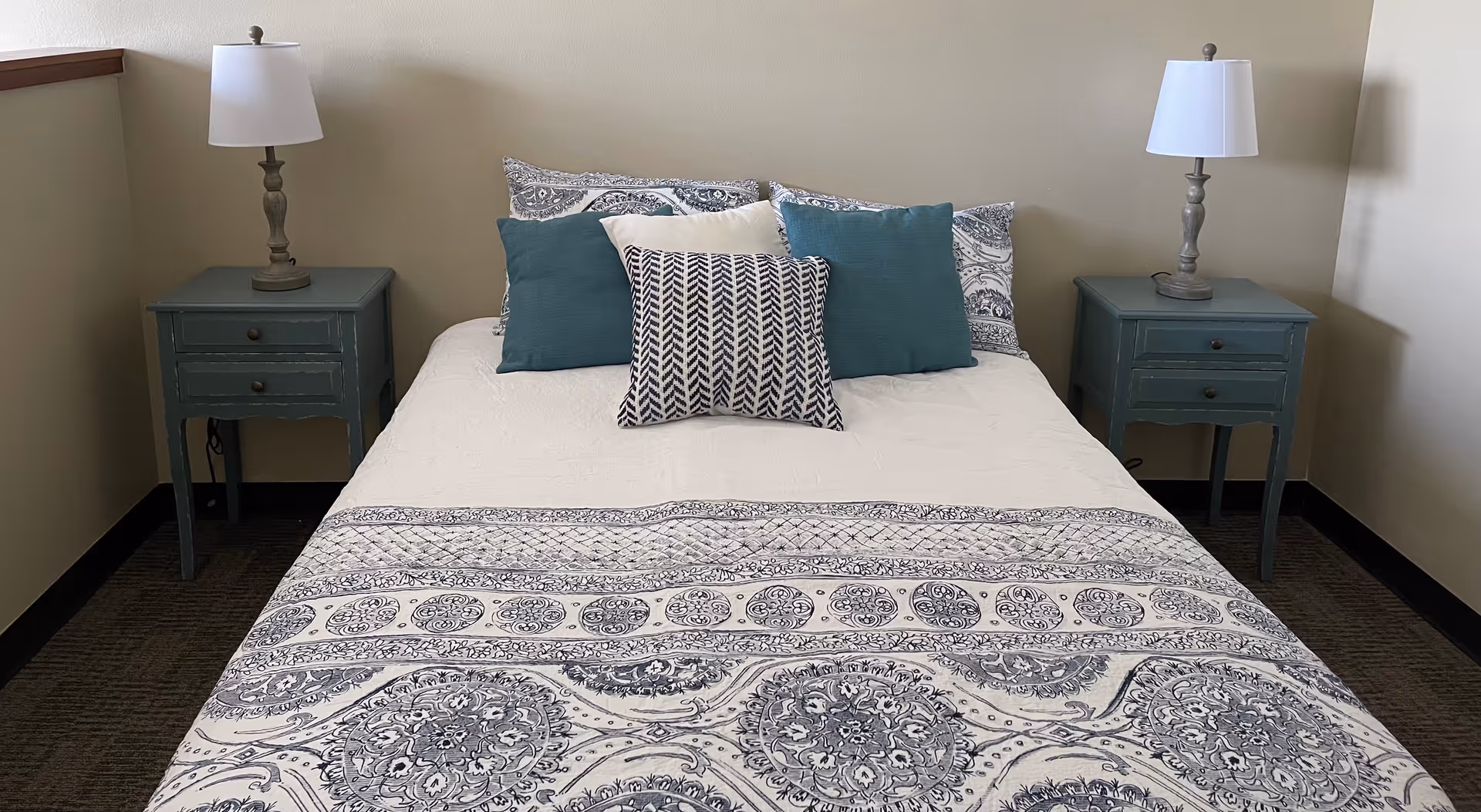 A neatly made bed with a white and gray patterned bedspread and multiple pillows in shades of blue and white. On each side of the bed is a small teal nightstand with two drawers, each topped with a lamp with a white lampshade. The room has beige walls and a carpeted floor.