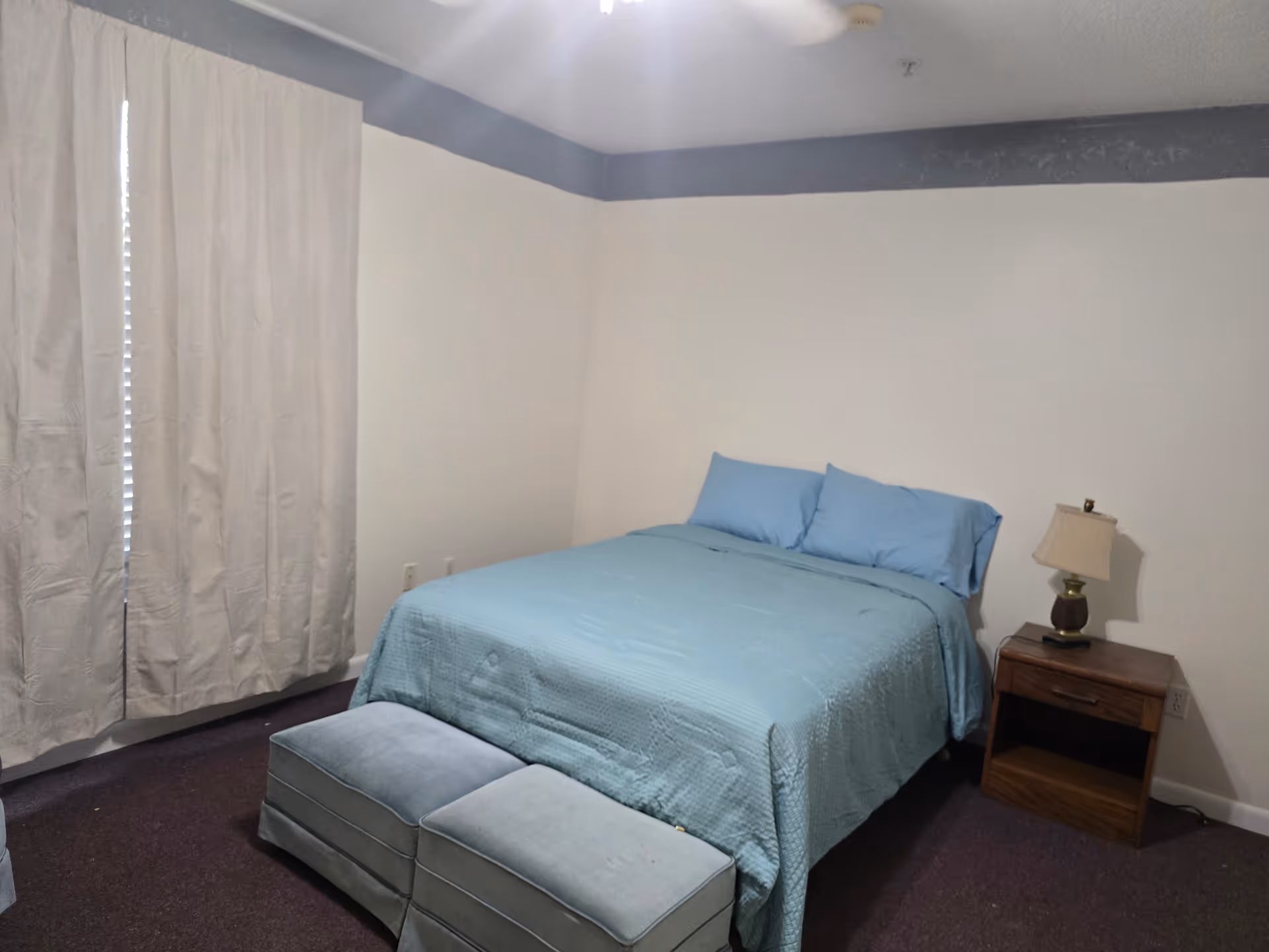 Simple bedroom in a senior living facility with a bed, nightstand, and curtains.