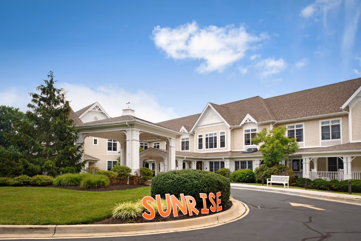 Exterior view of a senior living facility building with beige siding and white trim under a blue sky with some clouds. There is a covered entrance with white columns, well-maintained landscaping including bushes and a tree, and a curved driveway. Large orange letters spelling 'SUNRISE' are placed on the grass near the driveway.