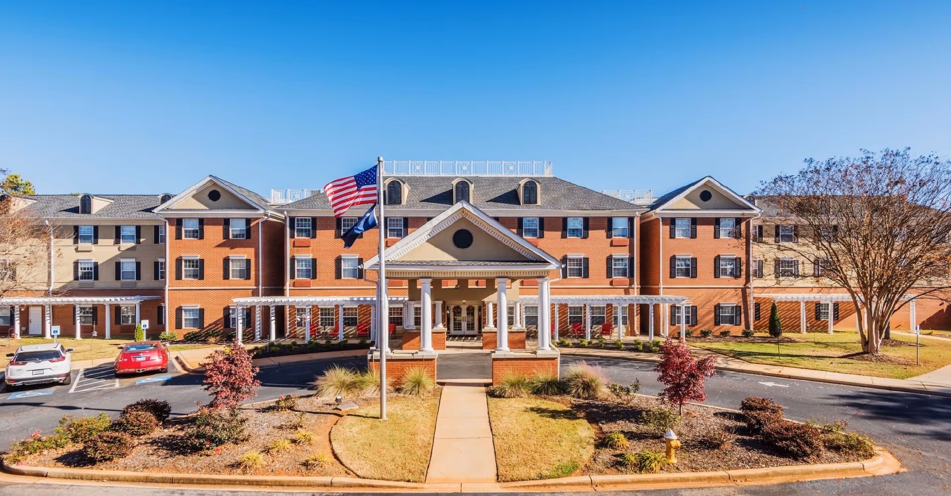 Front exterior of a three-story brick senior living building with a porte-cochère, flagpoles, parking area, and landscaped driveway.