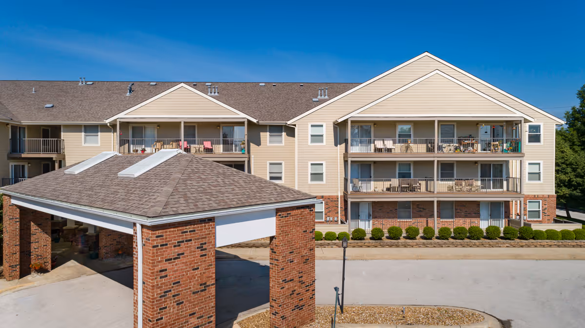 Exterior view of a multi-story assisted living facility building with beige siding and brick accents. The building has balconies with outdoor furniture and a covered entrance area with a peaked roof. The sky is clear and blue.
