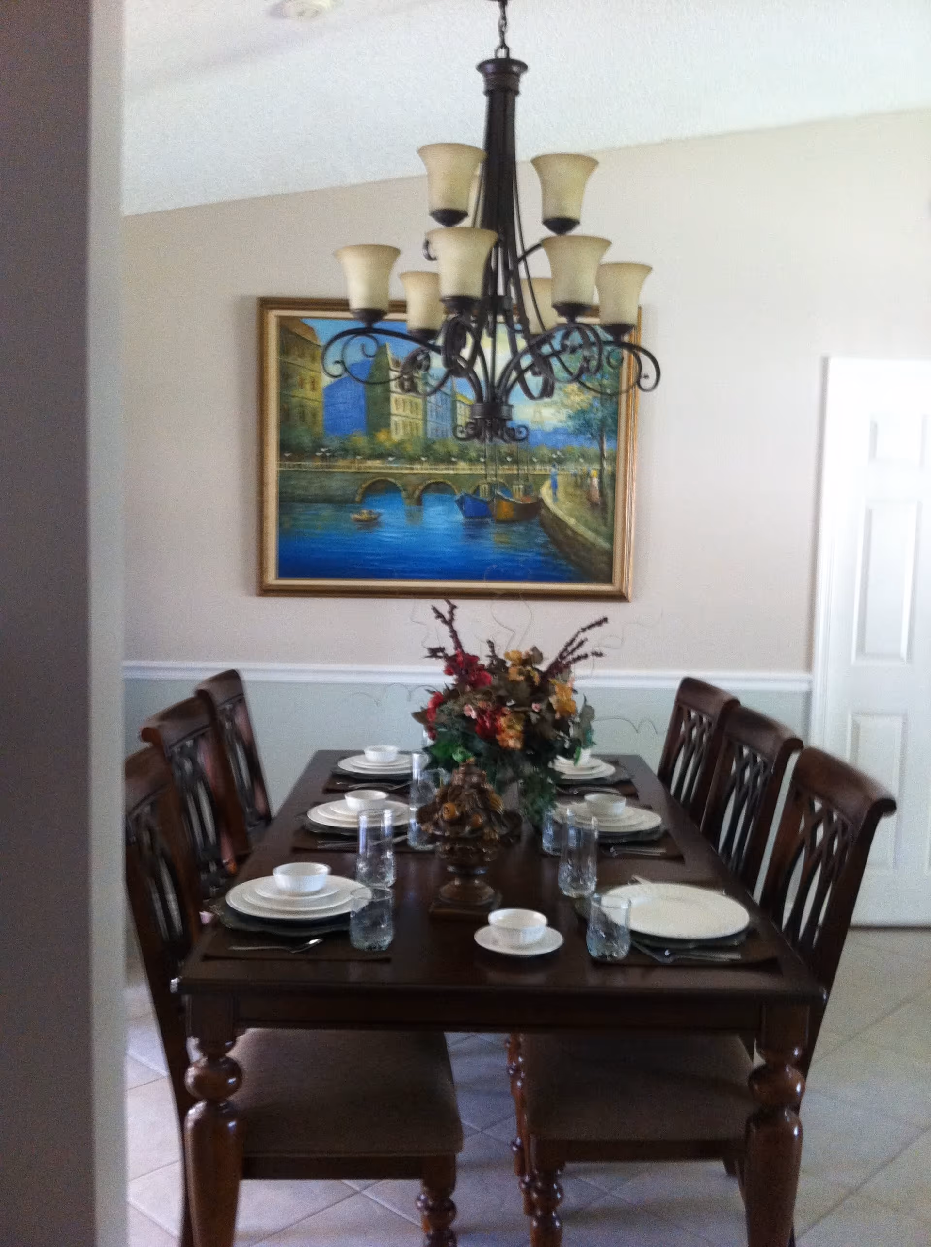 A dining room with a dark wooden table set for six people, including plates, bowls, glasses, and utensils. A floral centerpiece decorates the middle of the table. Above the table hangs a chandelier with multiple light fixtures. A framed painting of a riverside scene with buildings and boats is on the wall behind the table.