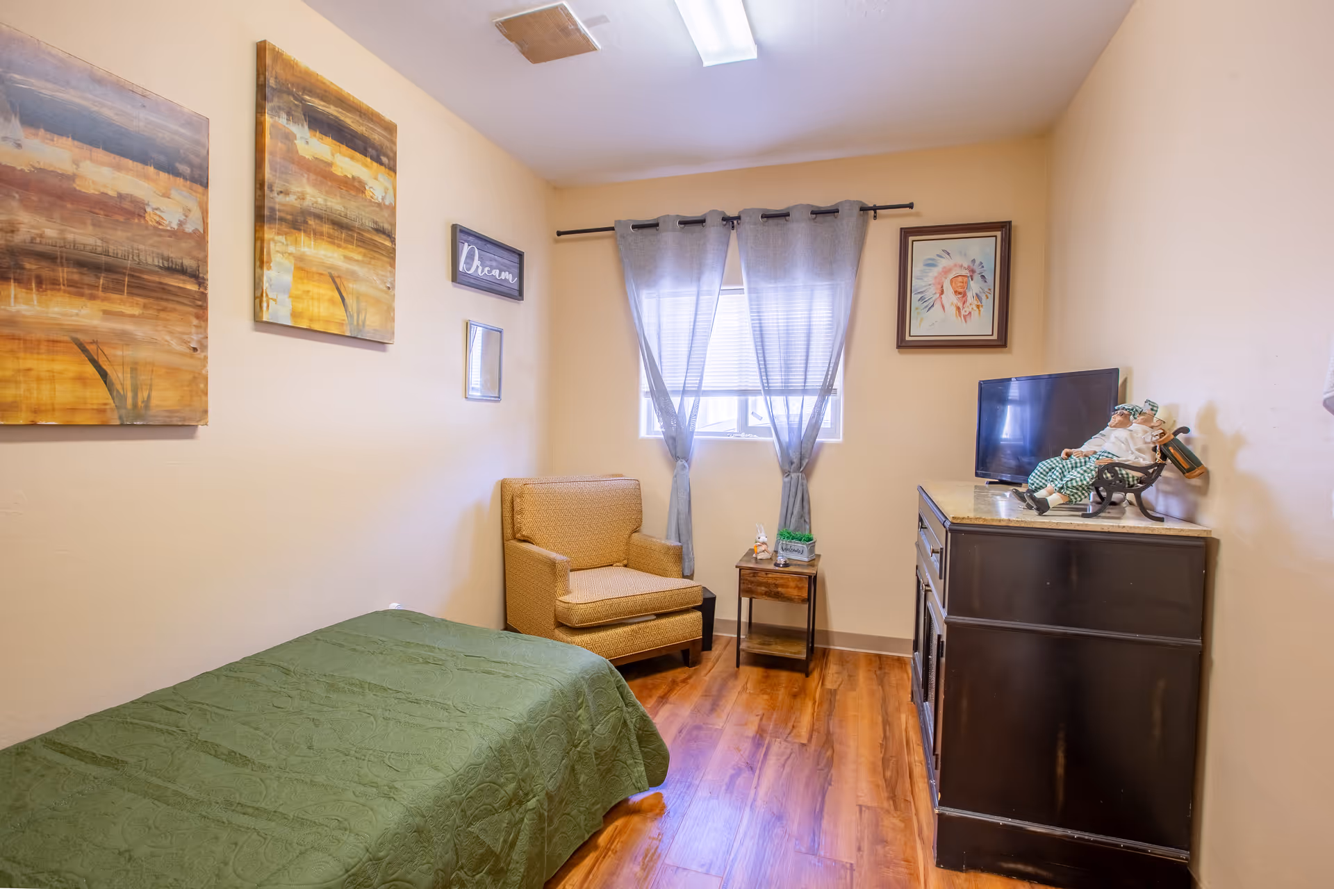 Cozy bedroom with a bed, chair, dresser, and window with sheer curtains in a facility.
