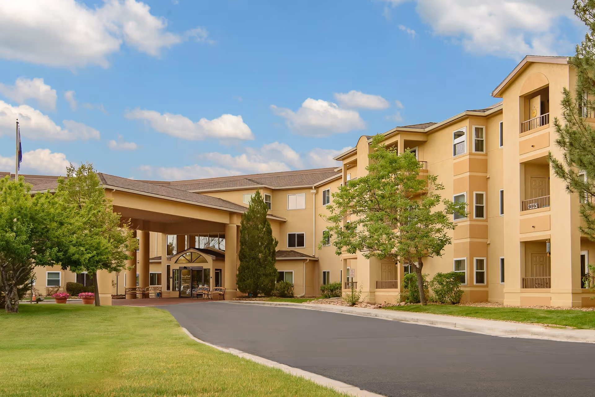 Front entrance and facade of a three-story beige senior living building with a covered porte-cochère, driveway, and trees.