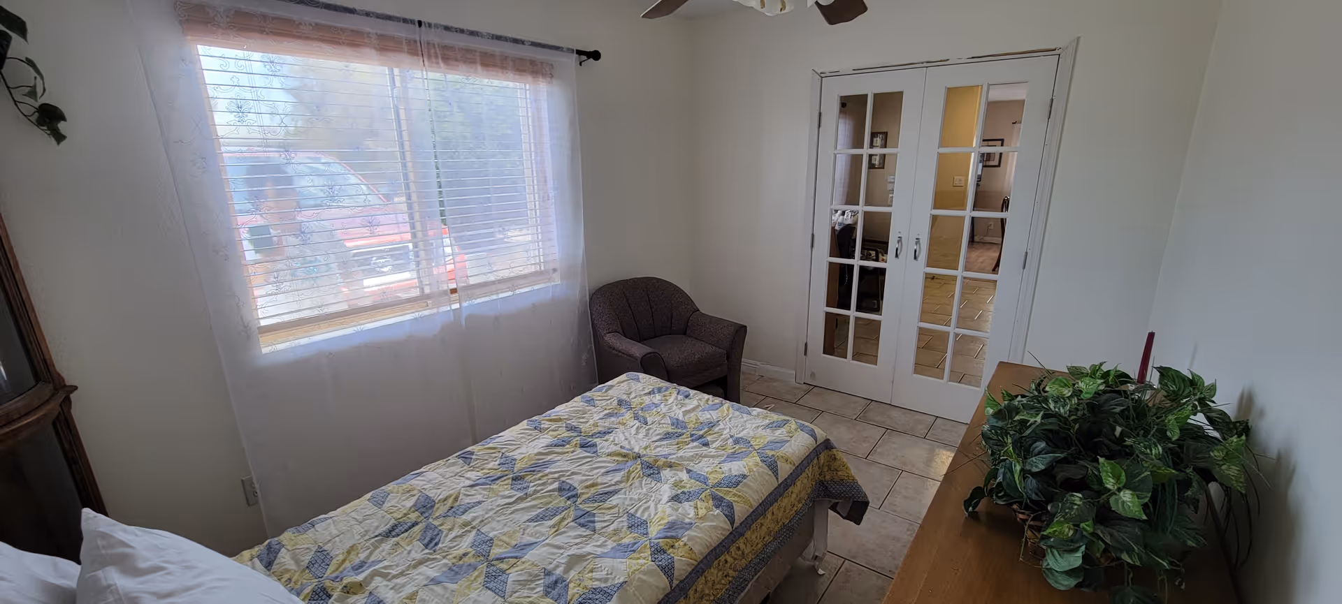 A small bedroom with a quilted bed, window, armchair, French doors, and a dresser with plants.