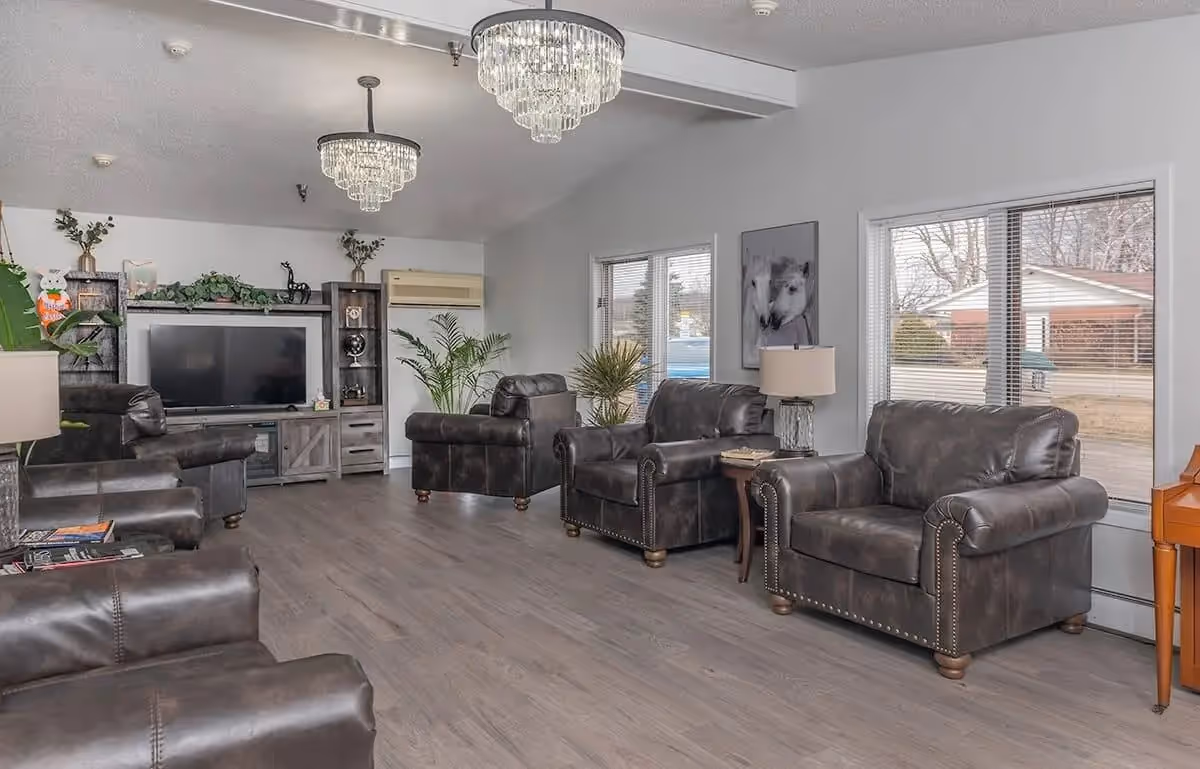 A spacious living room with multiple dark leather armchairs arranged around the room. The room features large windows with blinds, two crystal chandeliers hanging from the ceiling, a flat-screen TV on a wooden entertainment center, and various plants and decorative items. The floor is covered with light wood laminate.