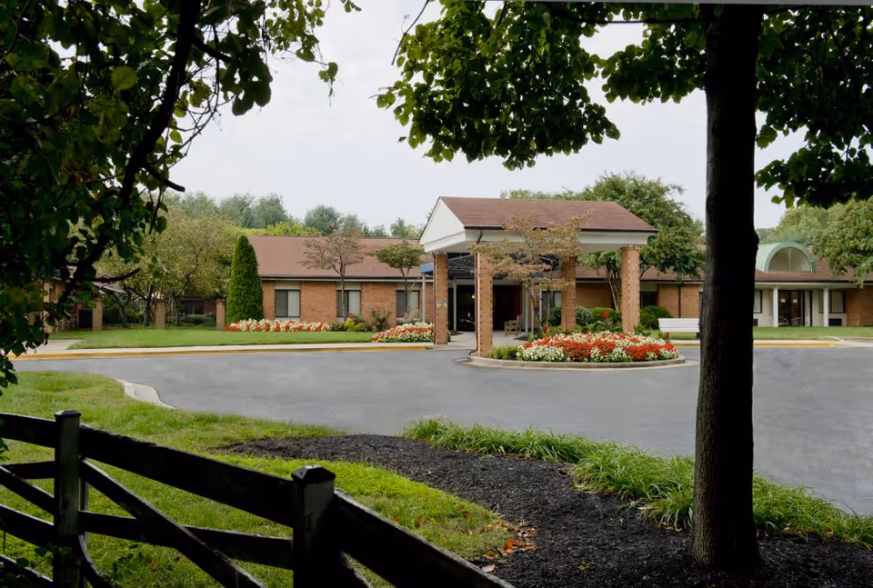 View of the front entrance of Pineview Manor Extended Care, showing a circular driveway with a landscaped flower bed in the center, surrounded by a brick building with a covered entryway. Trees and greenery frame the scene with a wooden fence in the foreground.