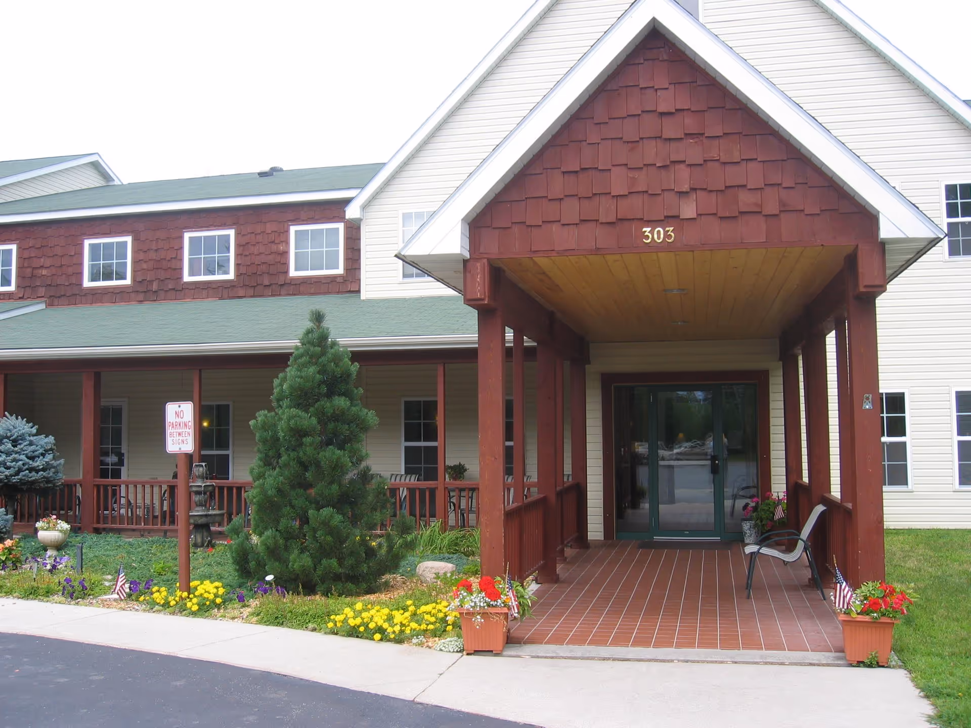 Entrance of a senior living facility with a covered porch supported by wooden pillars, a tiled floor, and a glass door. The building has beige siding with red shingles on the upper part and the number 303 above the entrance. There are flower pots with colorful flowers and small American flags on either side of the entrance, a small tree, and a 'No Parking Between Signs' sign near the sidewalk.