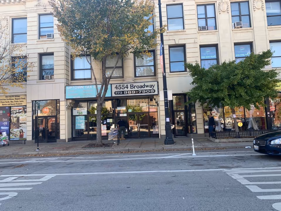 City storefronts along a street with pedestrians and trees in front of a multi-story building.