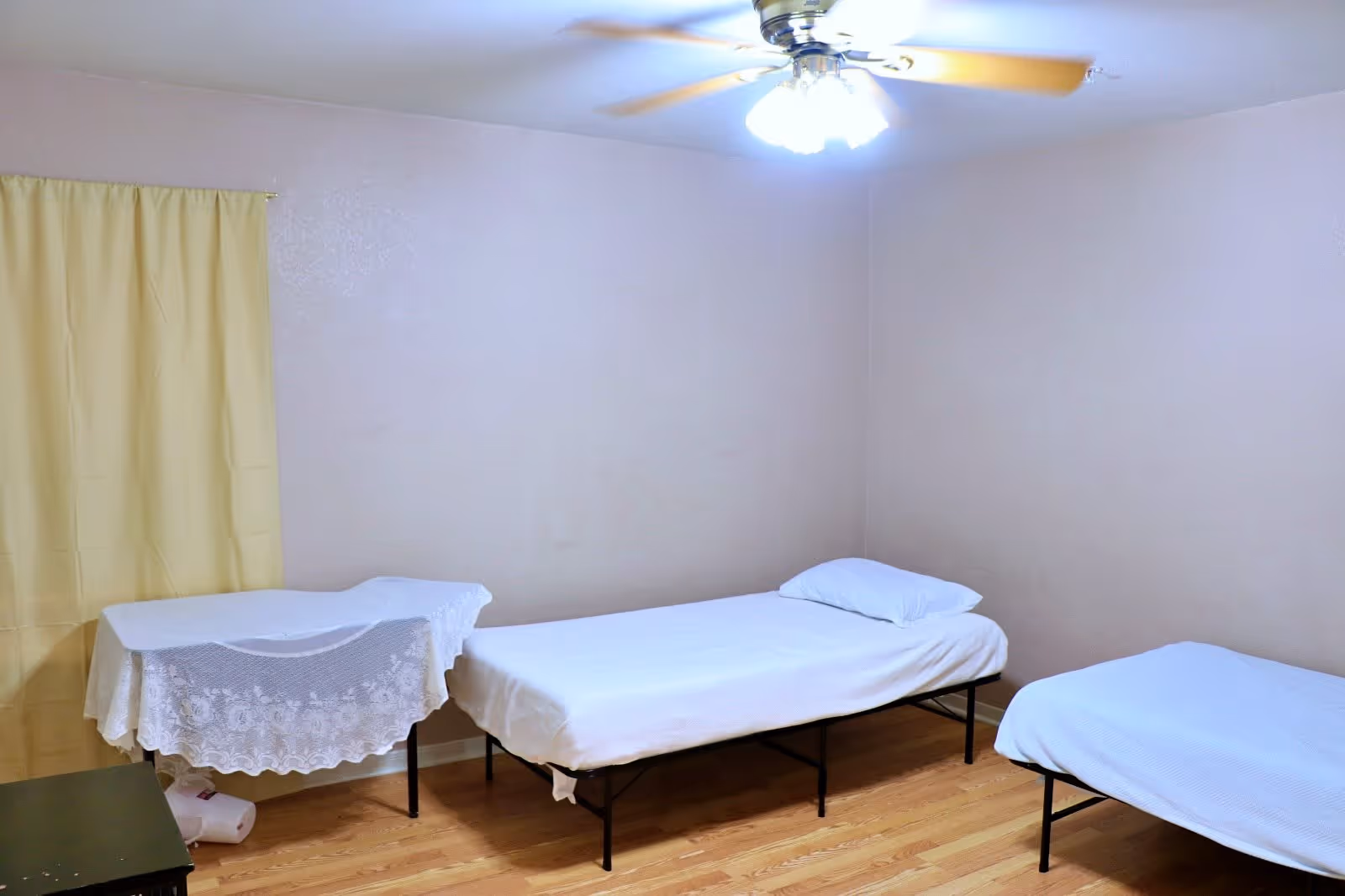 A simple bedroom with two single beds covered in white sheets and pillows. There is a small table with a white lace tablecloth near a window with a yellow curtain. The room has light-colored walls, a wooden floor, and a ceiling fan with lights.