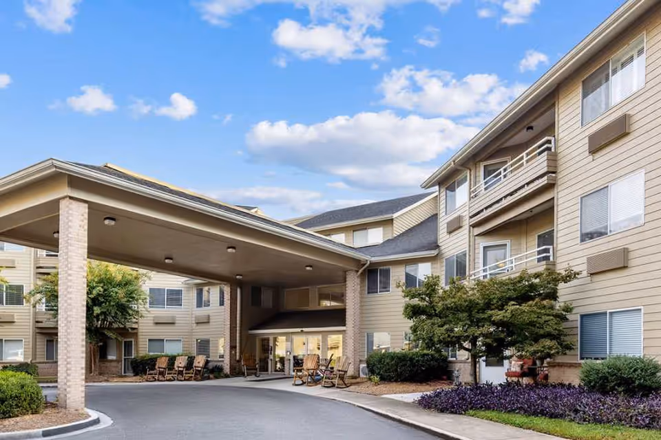 Exterior view of a multi-story senior living facility with beige siding and multiple windows. The entrance features a covered drop-off area supported by brick columns, with several wooden rocking chairs arranged near the entrance. There are landscaped bushes and a small tree near the building, under a partly cloudy blue sky.