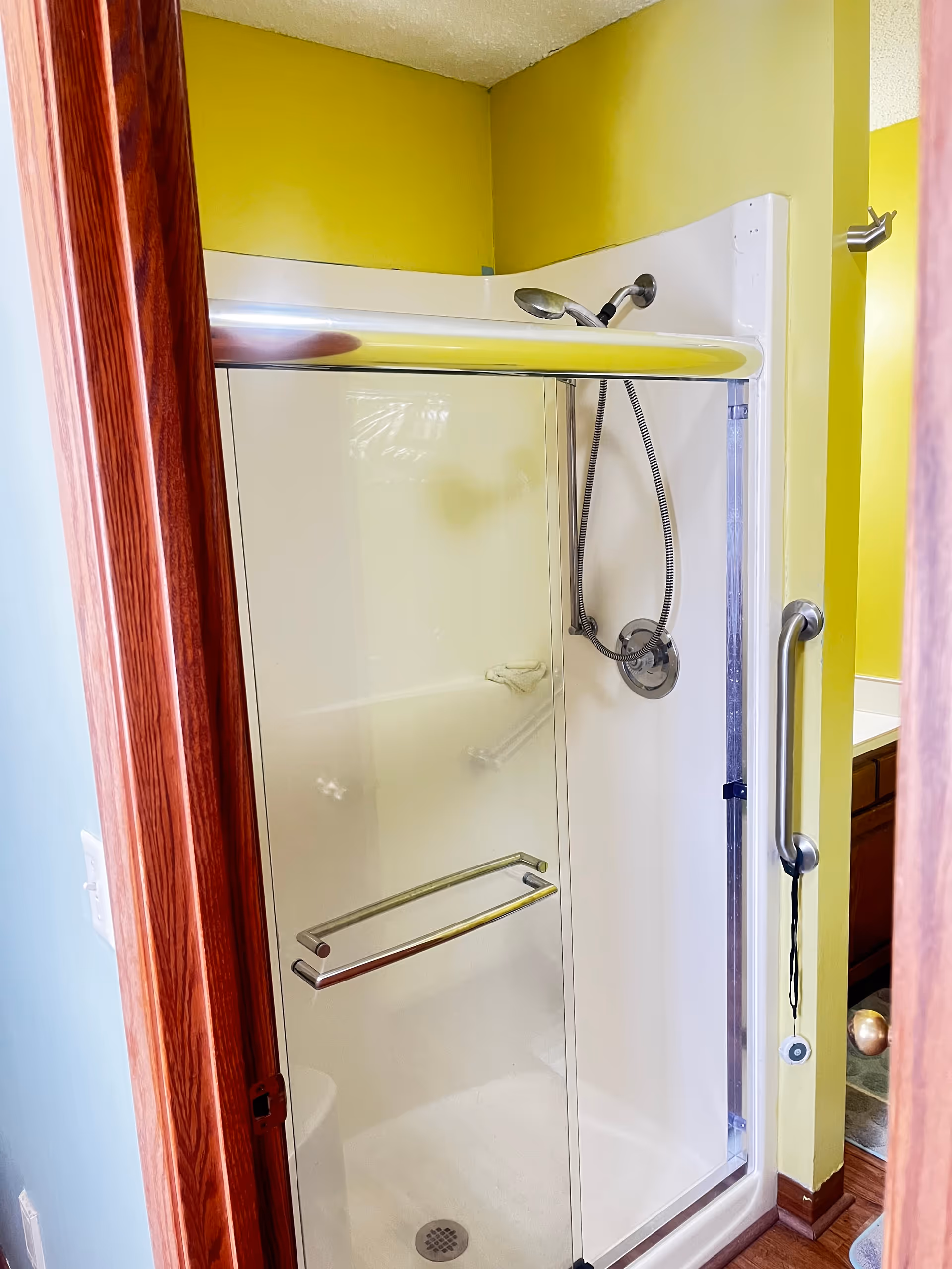 Accessible shower with glass doors, grab bar, and handheld shower in a yellow-walled bathroom.