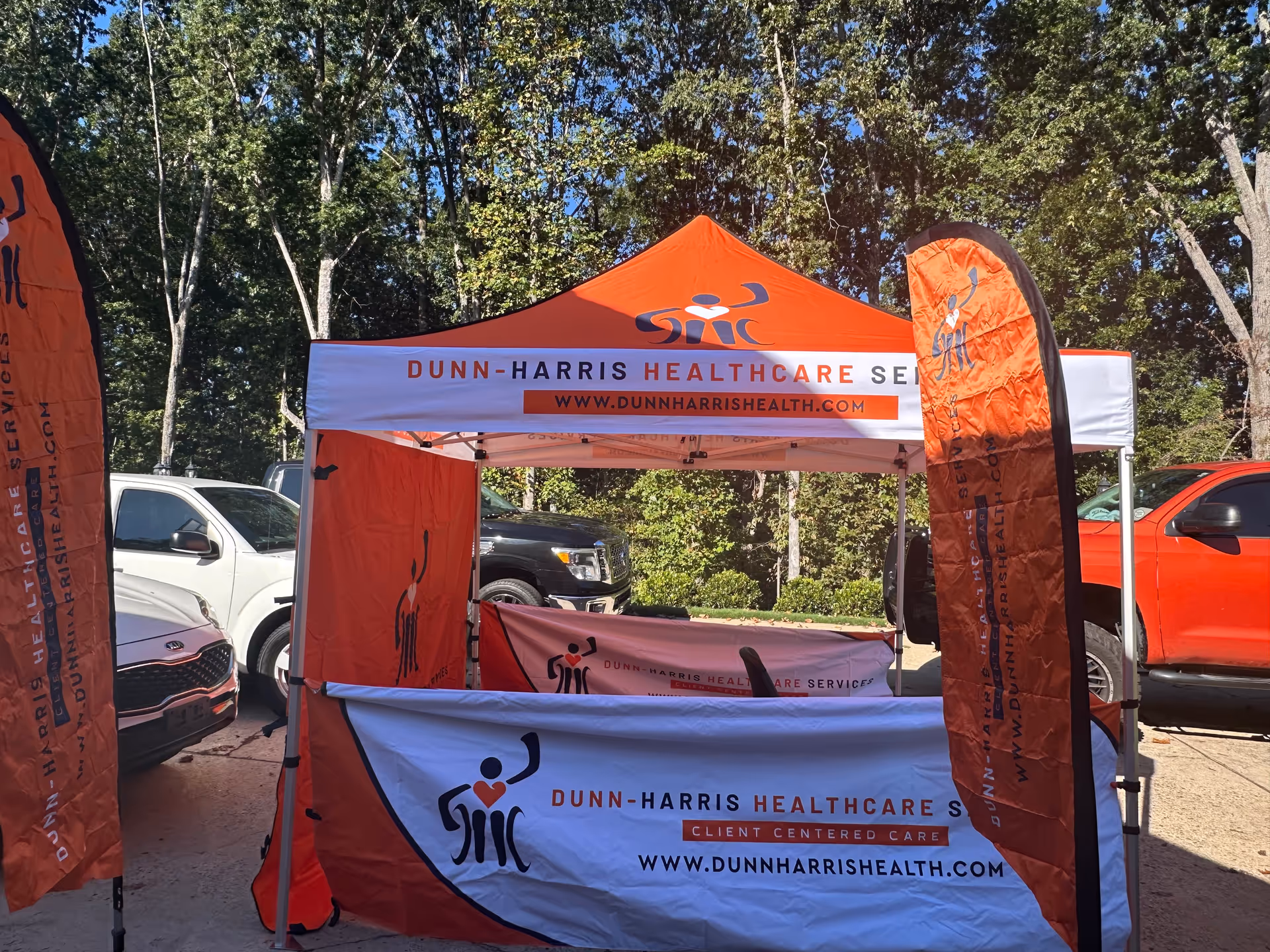 Orange Dunn-Harris Healthcare tent and banners set up outdoors beside parked cars.
