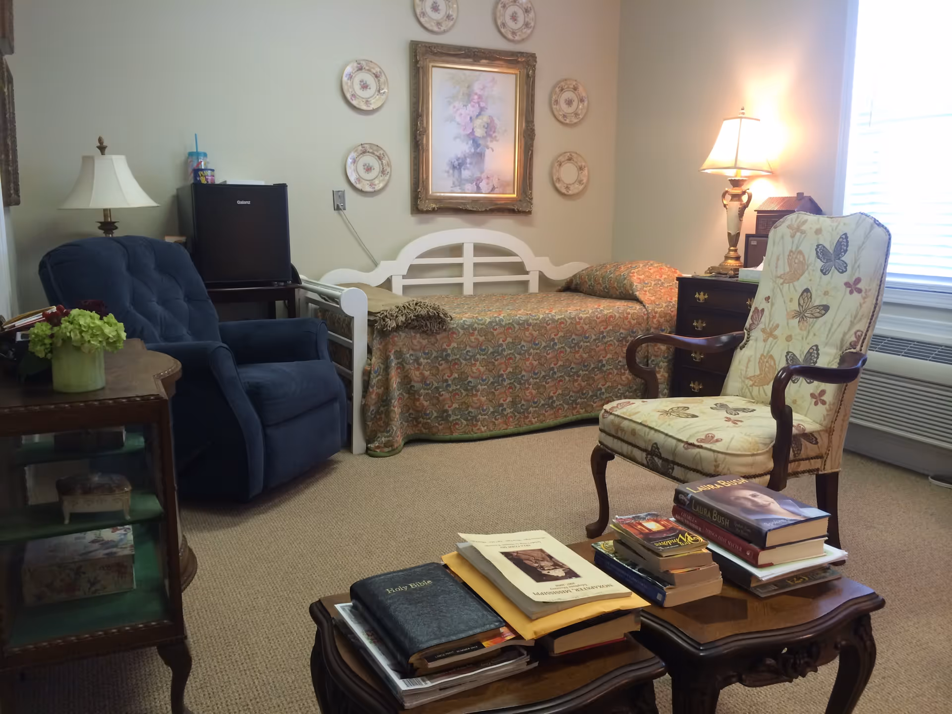 A cozy room with a single bed covered in a patterned bedspread, a blue armchair, a floral upholstered chair, and two wooden tables stacked with books and papers. The walls are decorated with a framed floral painting surrounded by decorative plates. A lamp is lit on a dark wooden dresser near a window with blinds.