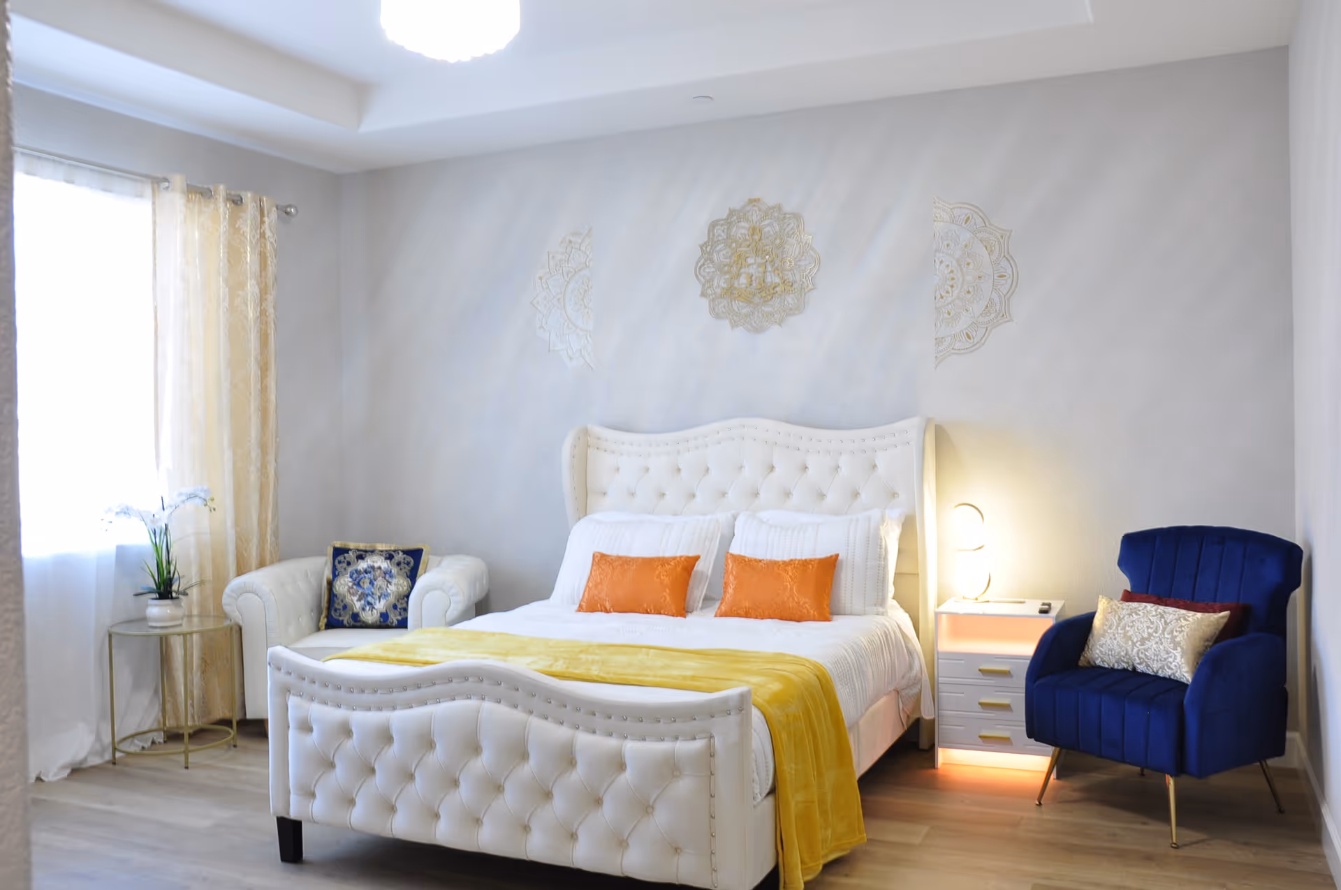 Bright bedroom with a tufted white bed, yellow throw and two colorful accent chairs.