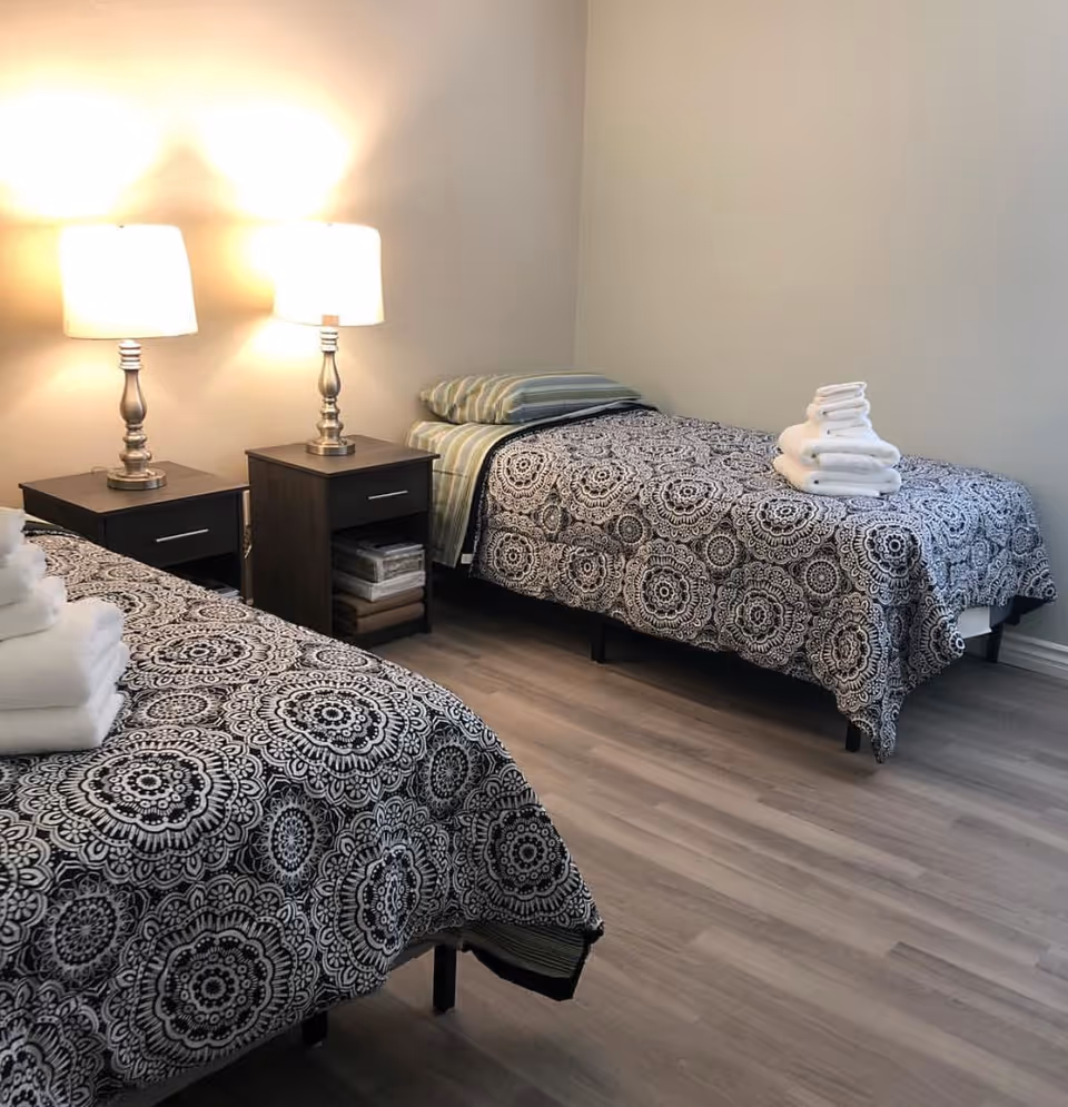A simple bedroom with two twin beds covered in black-and-white patterned bedspreads, a shared nightstand with two table lamps, and folded towels on the beds.