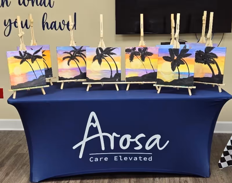 Display table with palm-tree sunset paintings on easels inside Arosa senior living facility.