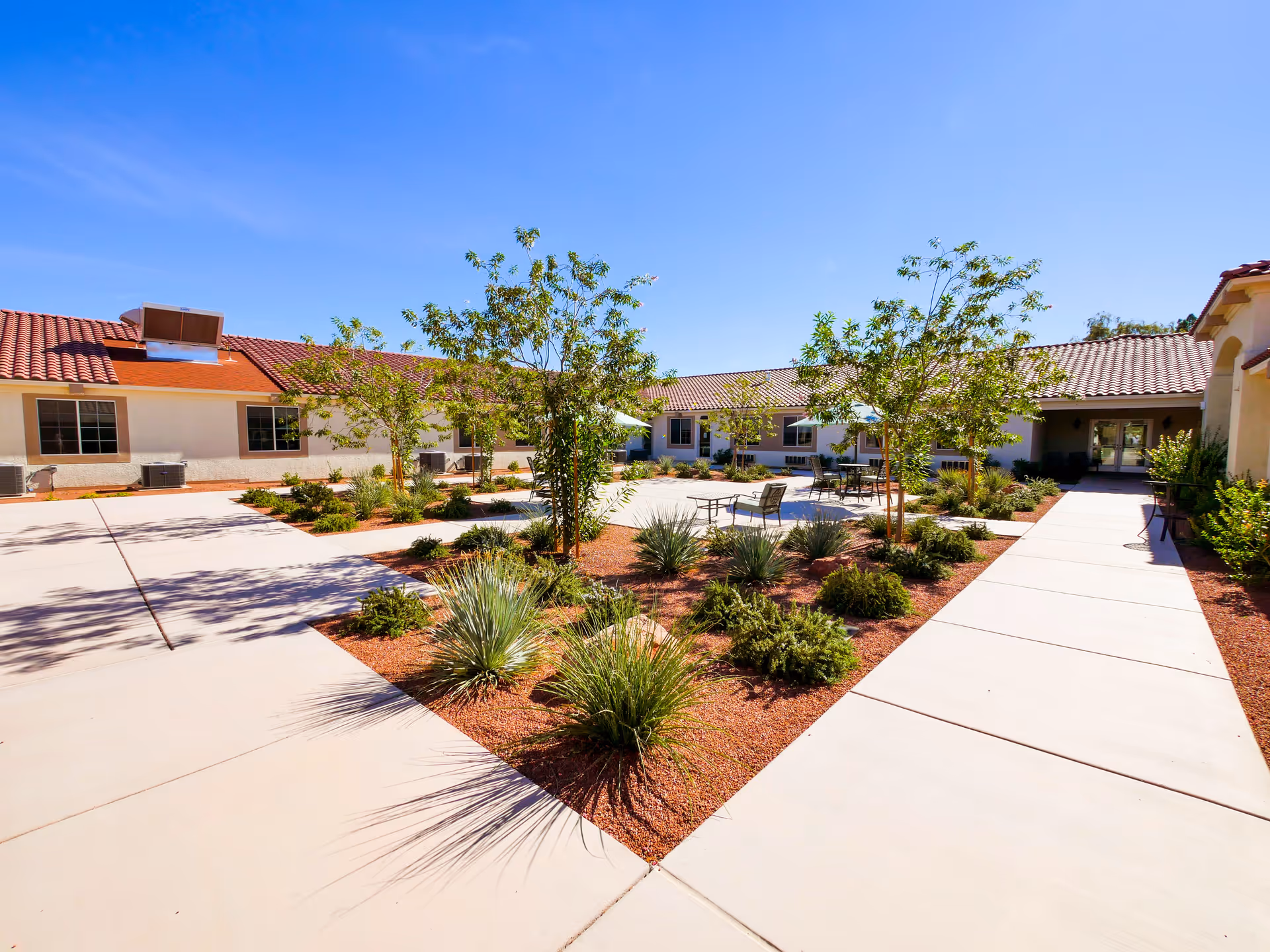 Outdoor courtyard area at Silver Hills Health Care Center featuring paved walkways, landscaped garden beds with small trees and shrubs, and several seating areas with tables and chairs under umbrellas, all under a clear blue sky.