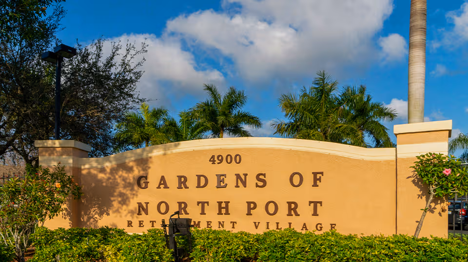 Entrance sign for Gardens of North Port Retirement Village with palm trees and blue sky.