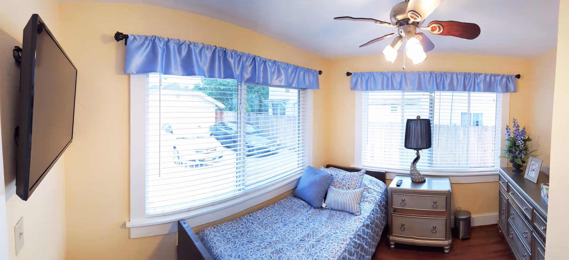 A small bedroom with a single bed covered in blue patterned bedding and three pillows. There are two windows with blue valances letting in natural light. A ceiling fan with lights is on the ceiling. Next to the bed is a silver nightstand with a black lamp and a remote control. A silver dresser with a flower arrangement and a framed picture is against the wall. A flat-screen TV is mounted on the wall opposite the bed.