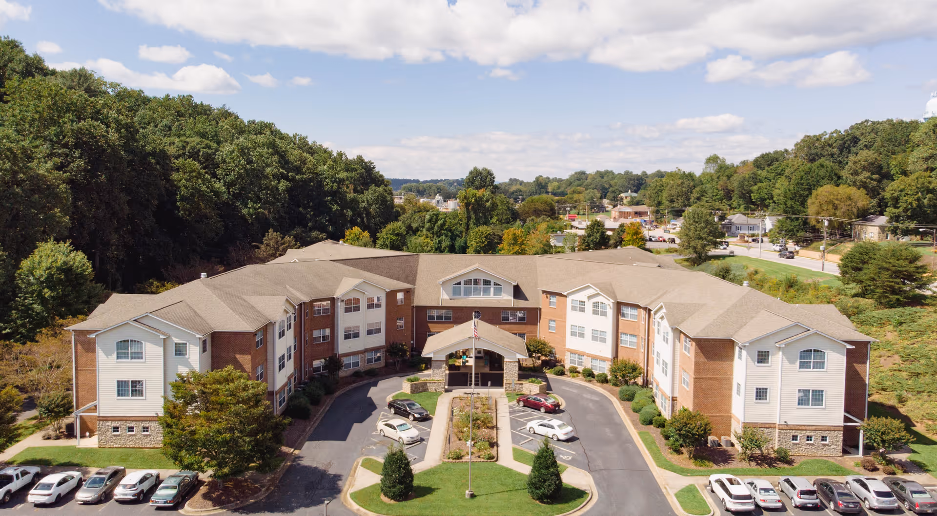 Aerial view of Rose Glen Village showing a three-story U-shaped brick-and-siding senior living building with entrance drive, parking, and surrounding trees.