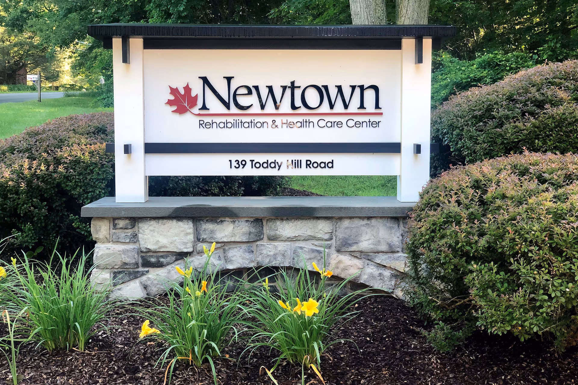 Entrance sign reading "Newtown Rehabilitation & Health Care Center" mounted on a stone base surrounded by shrubs and yellow flowers.