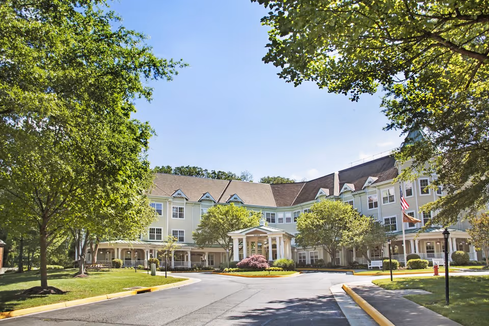 Front view of a multi-story senior living facility with a covered entrance, circular driveway, and landscaped trees and lawn.