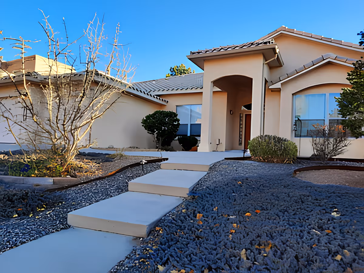 Front exterior view of a single-story building with a beige facade, tiled roof, and a covered entrance supported by columns. There is a paved walkway leading up to the entrance, surrounded by landscaped areas with bushes and leafless trees under a clear blue sky.