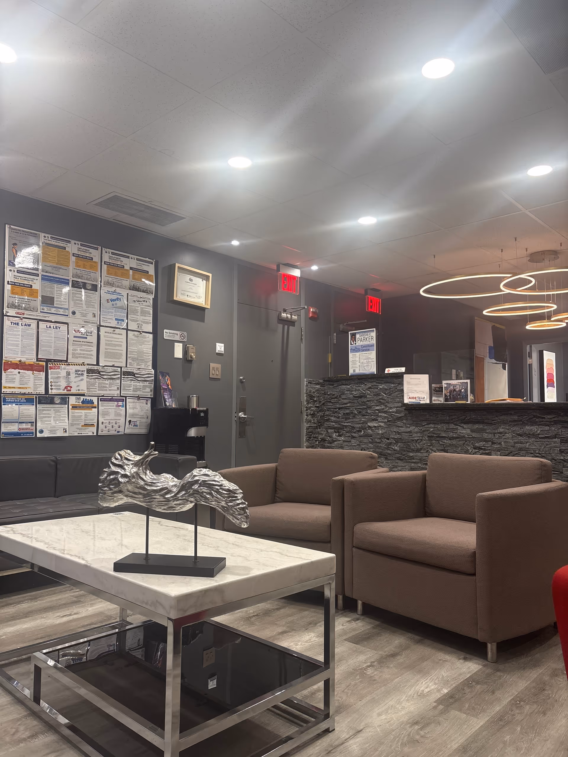 Modern waiting area with brown chairs, marble coffee table, stone reception desk, and posted notices nearby.