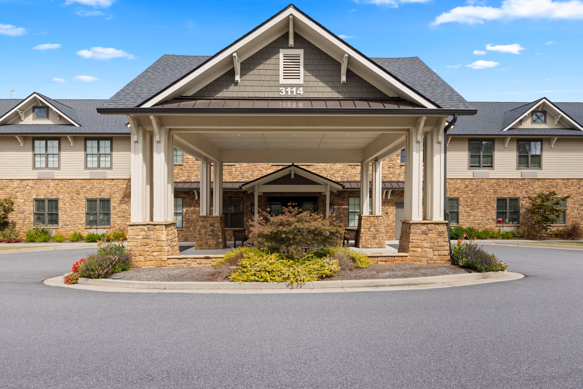 Front exterior view of a senior living facility building with a covered entrance supported by stone pillars, landscaped with bushes and flowers, under a blue sky with some clouds.