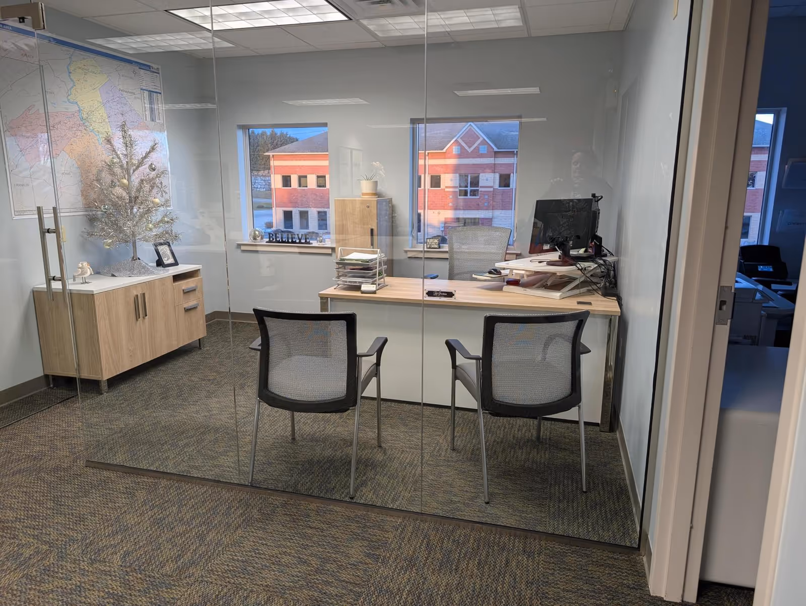 Glass-walled office with a desk, two chairs, computer, and a large map on the wall.