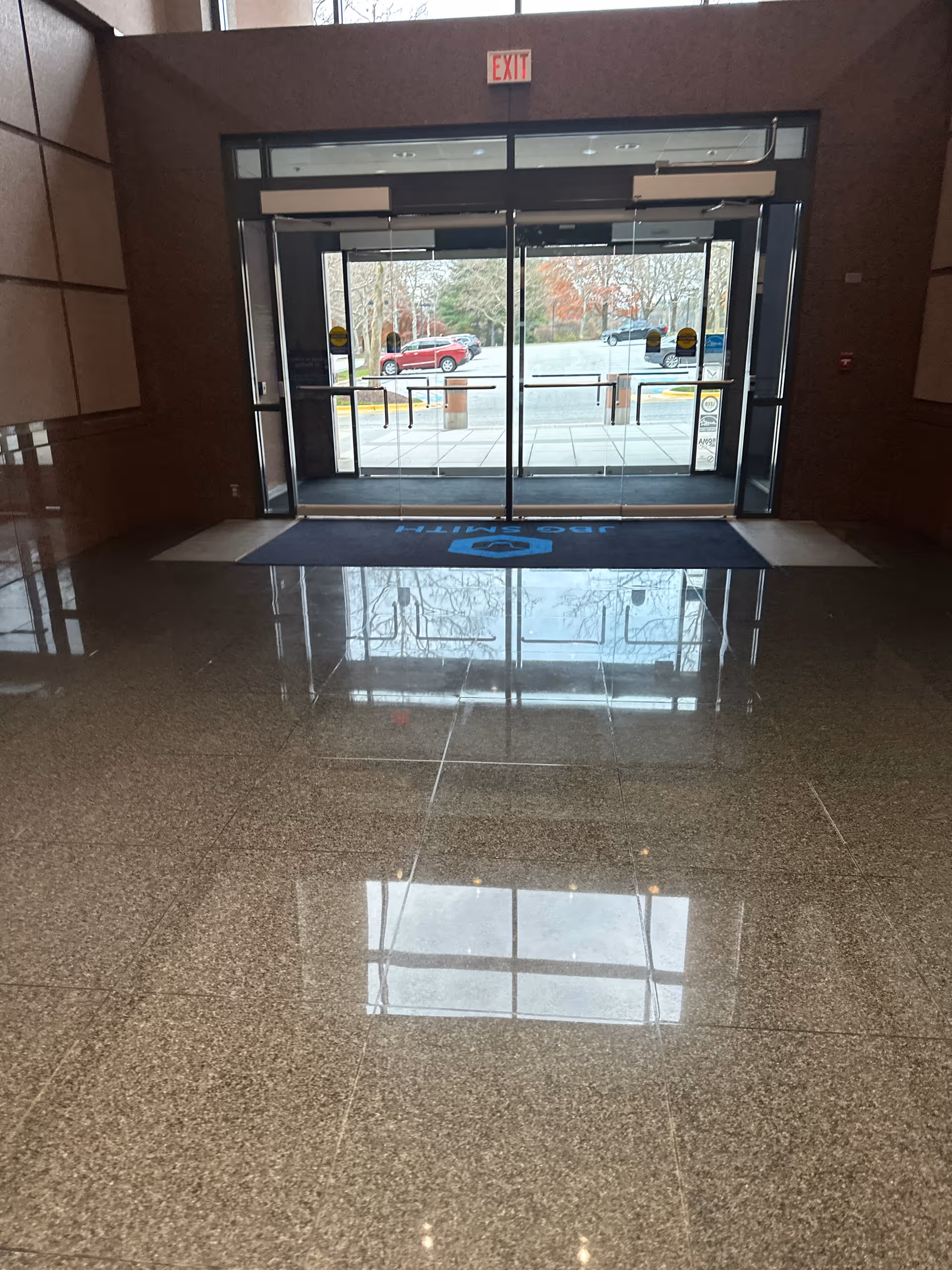 Interior lobby with glass automatic doors, exit sign, and polished tile floor reflecting outside trees.