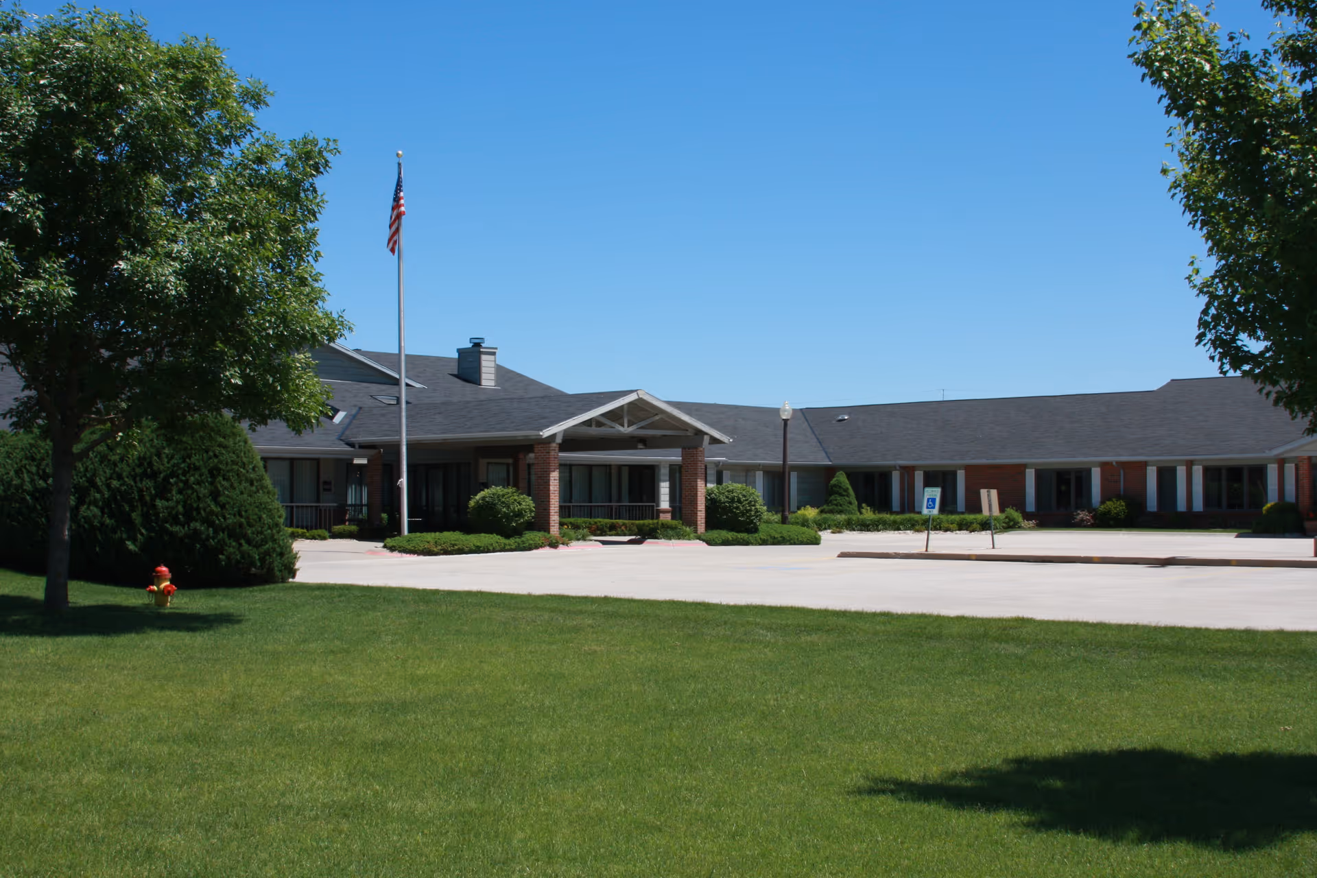 Exterior view of Riverside Lodge Retirement Community building with a large green lawn in the foreground, trees on either side, an American flag on a flagpole near the entrance, and a clear blue sky above.