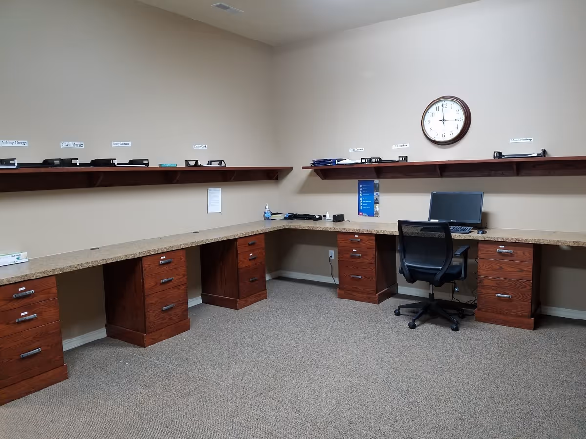 Empty office workspace with L-shaped desks, drawers, computer, and wall clock.