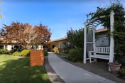 Curved walkway leads to a beige senior living building with a white porch bench and trees.